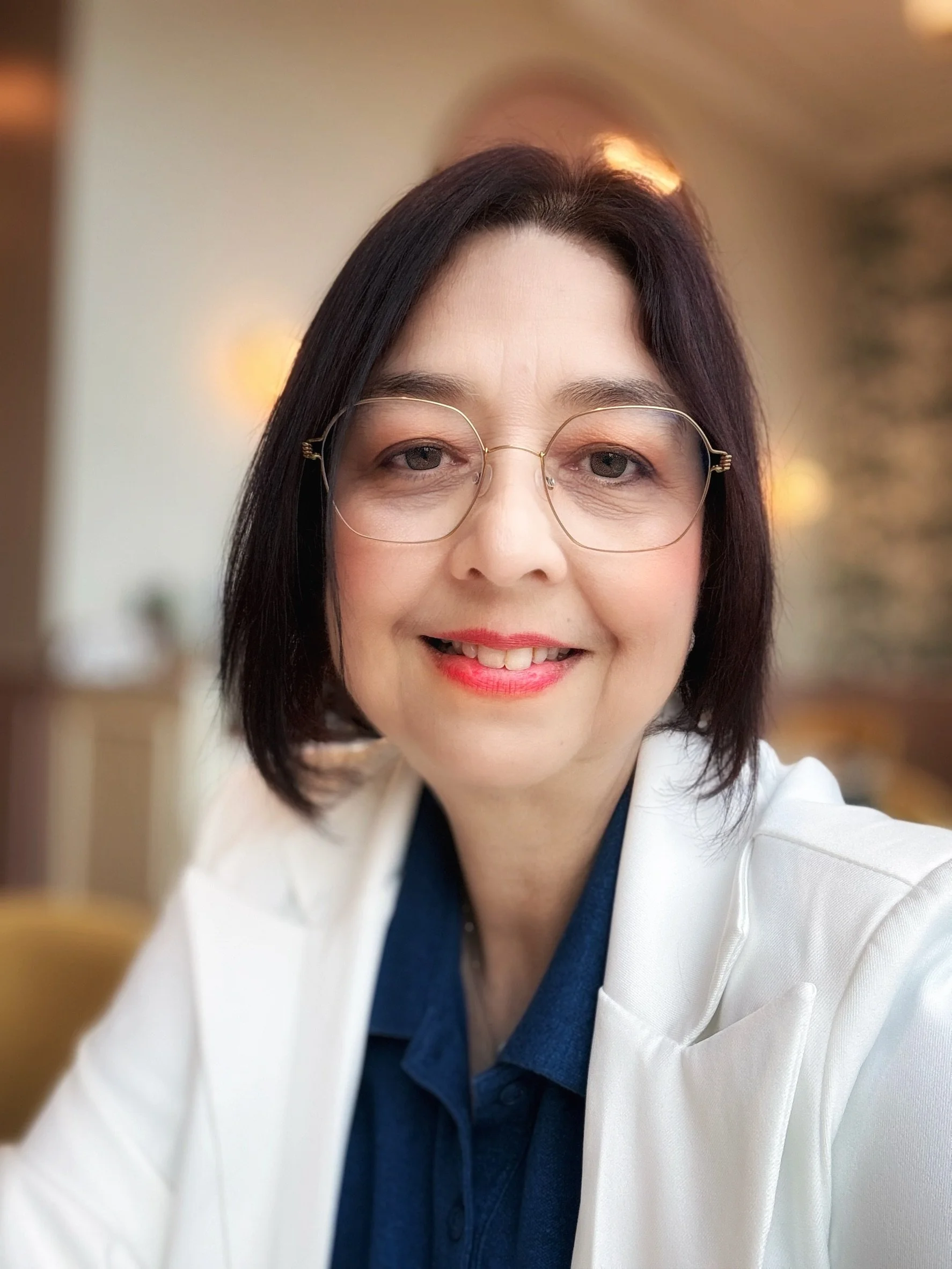Close-up of a woman with black hair and glasses smiling, wearing a white coat and dark top, with a blurred indoor background.