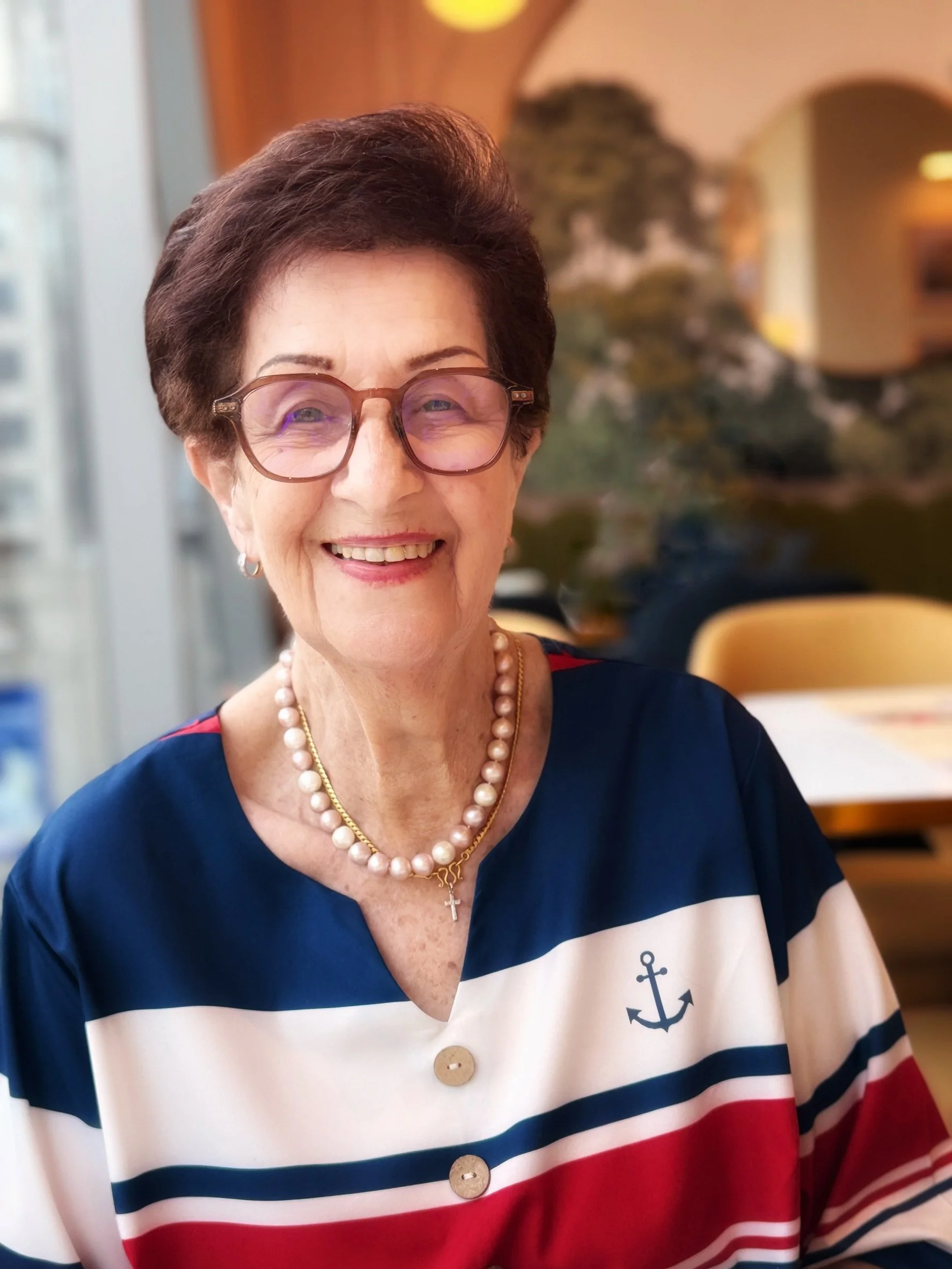 A smiling elderly woman with short dark hair and glasses, wearing a navy, white, red, and light pink striped sailor-style shirt with an anchor emblem, a pearl necklace, and a cross necklace. The background is blurred with warm tones and an arched doorway.