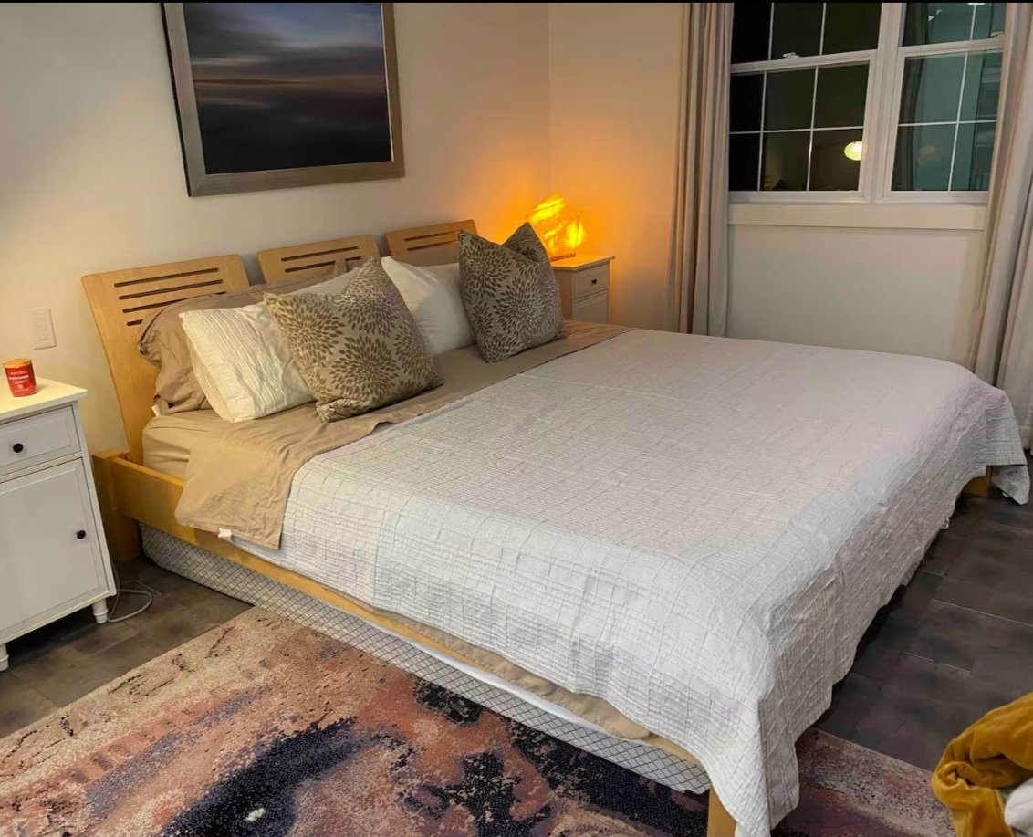 Bedroom with a neatly made bed featuring white and beige bedding, decorative pillows, wooden headboard, and bedside tables with lamps. A landscape picture hangs on the wall, and a window with curtains is on the right. A colorful rug covers the floor.