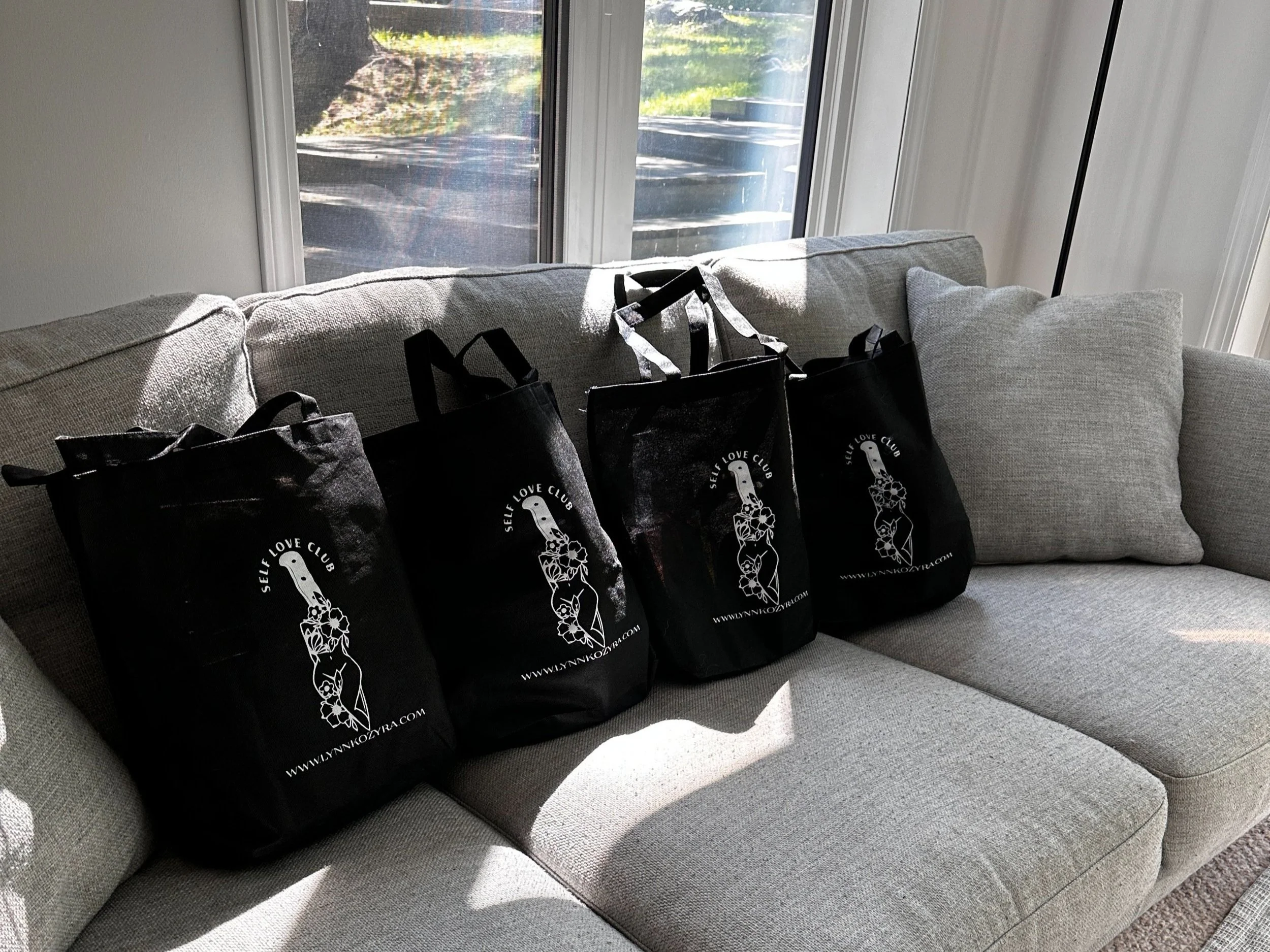 Four black tote bags with "Self Love Club" text and illustrations, placed on a gray couch near a window, with sunlight coming in.