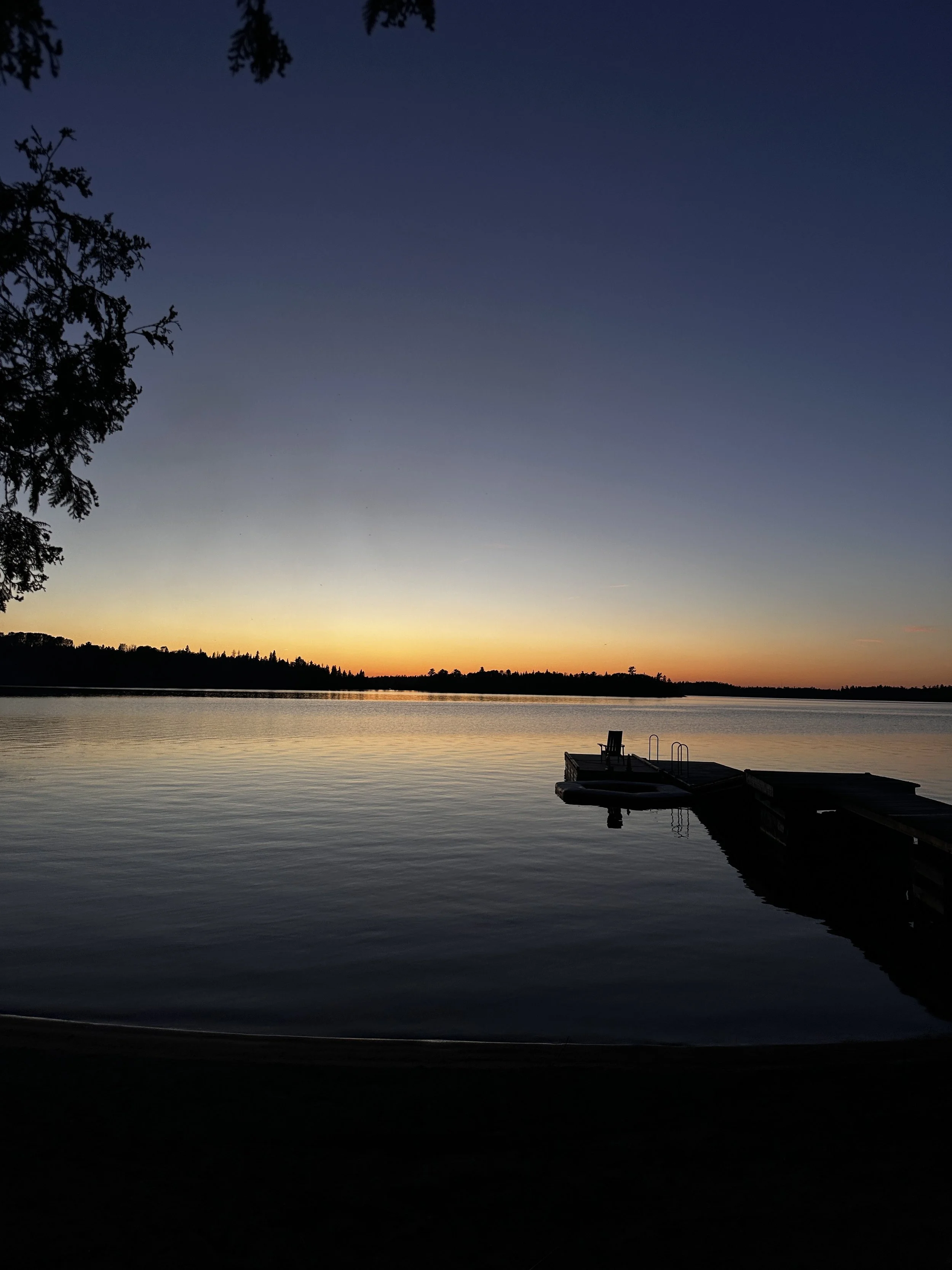 Sunset over a calm lake with a dock and chair, silhouetted trees on the horizon.
