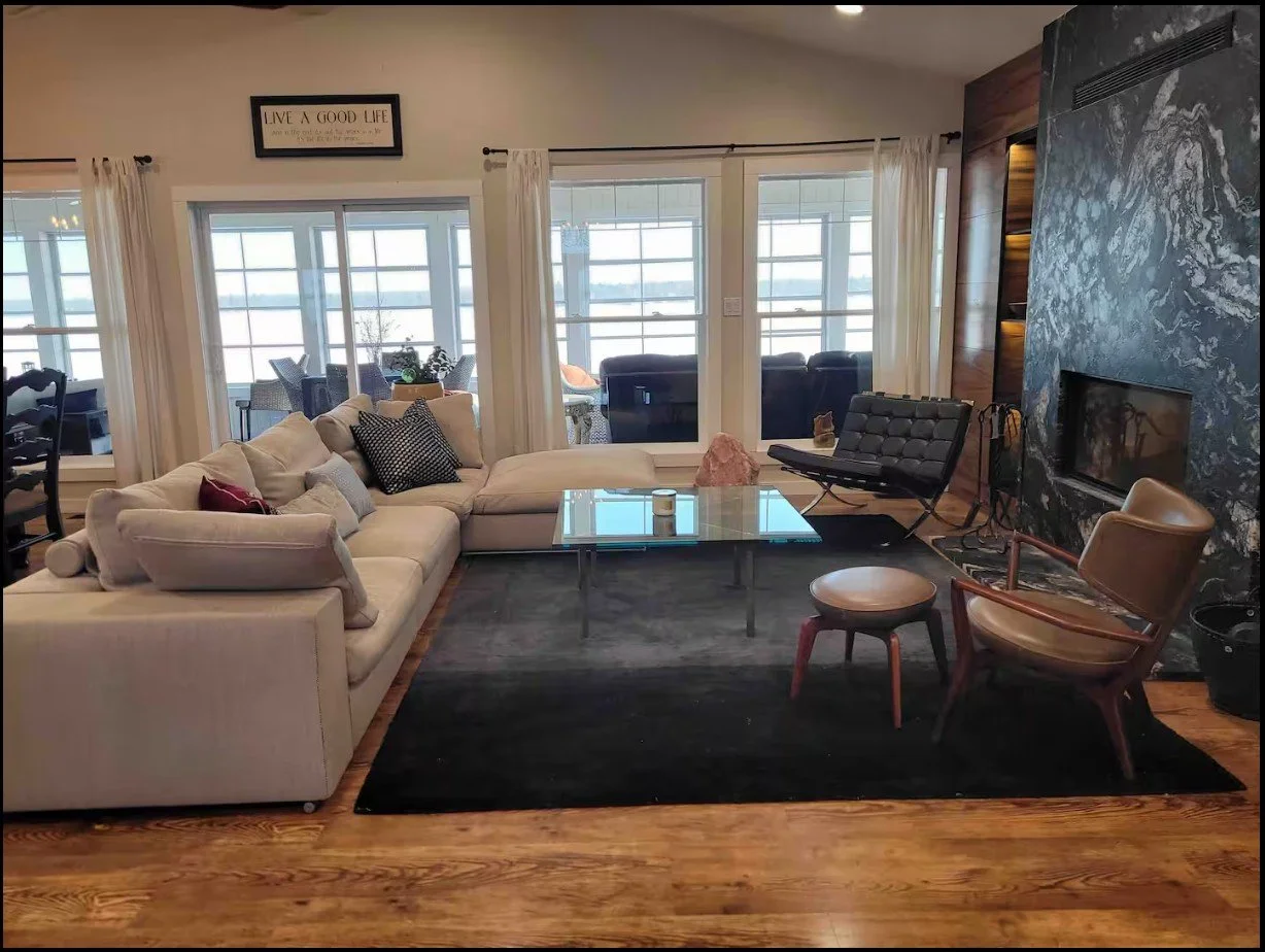 Contemporary living room with a large beige sectional sofa, glass coffee table, modern black chair, leather armchair, and a marble fireplace. Large windows provide natural light, and a "Live a Good Life" sign hangs above. A small outdoor patio is vis