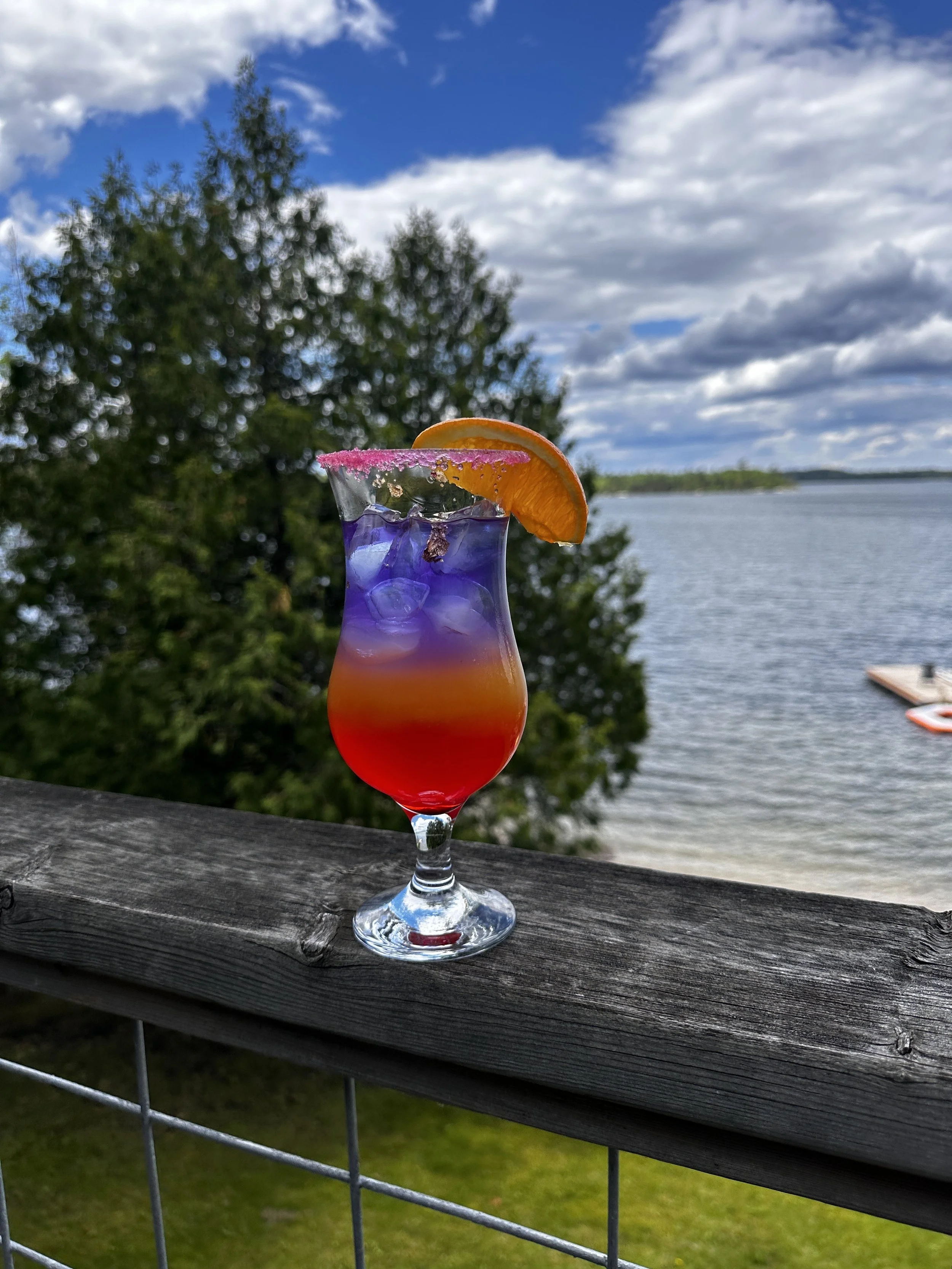 A colorful layered cocktail in a glass with pink sugar rim, topped with an orange slice, placed on a wooden railing overlooking a lake and trees under a partly cloudy sky.