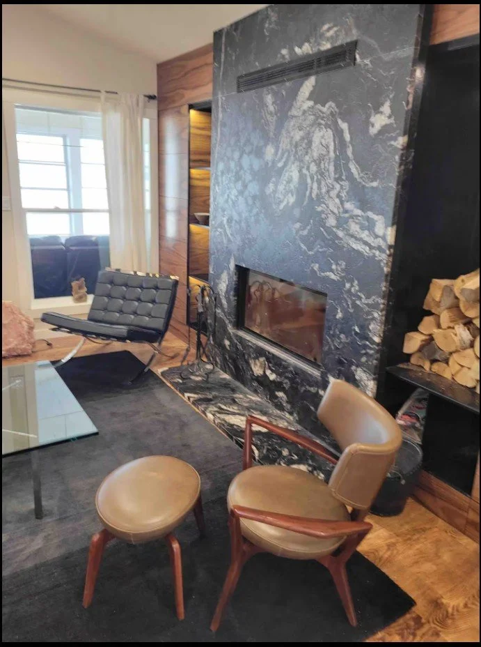 Modern living room with marble fireplace, chair, ottoman, and lounge chair. Glass coffee table and stacked firewood visible.