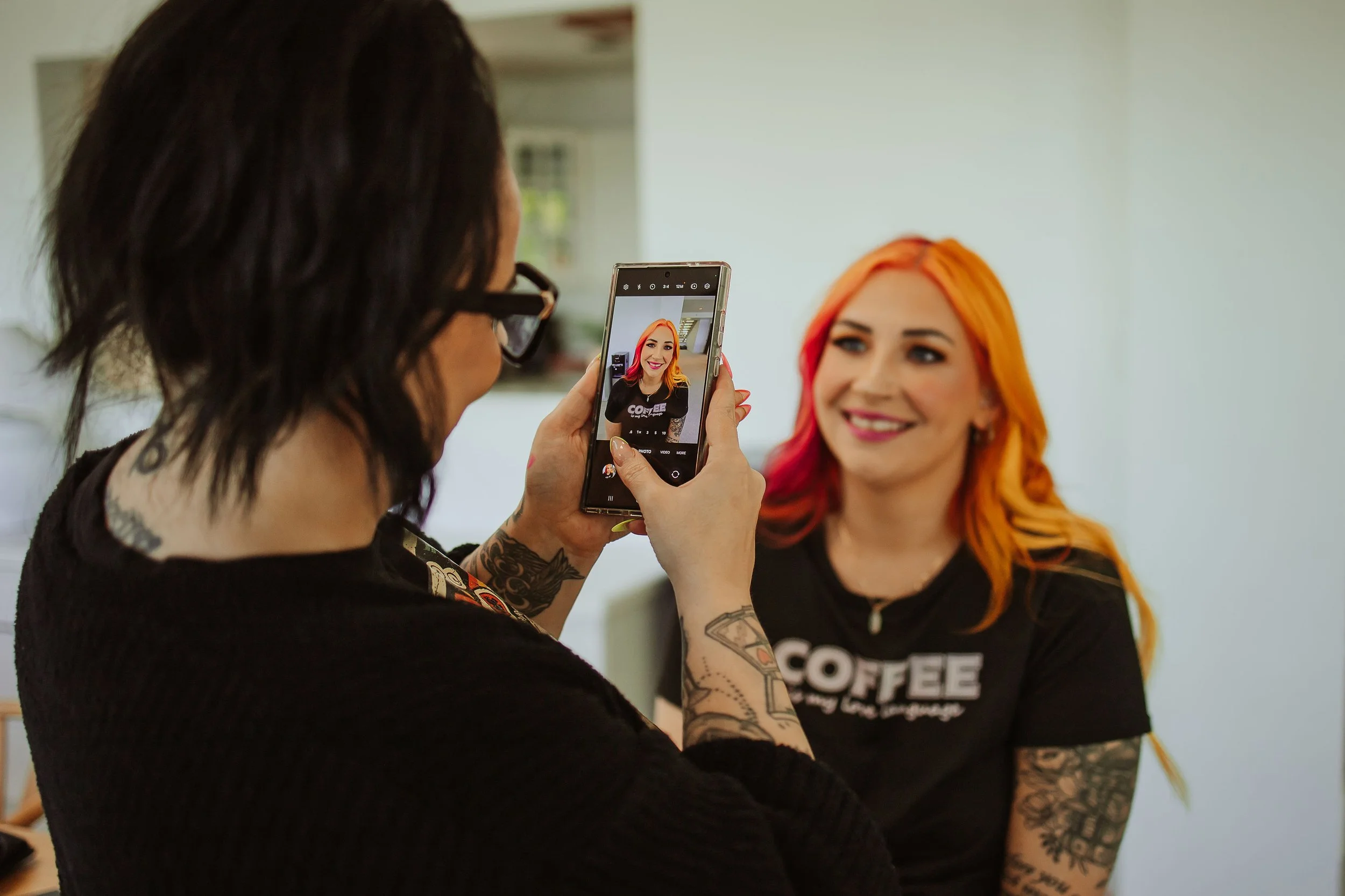 A person with black hair and tattoos taking a photo of a smiling person with colorful hair using a smartphone indoors.