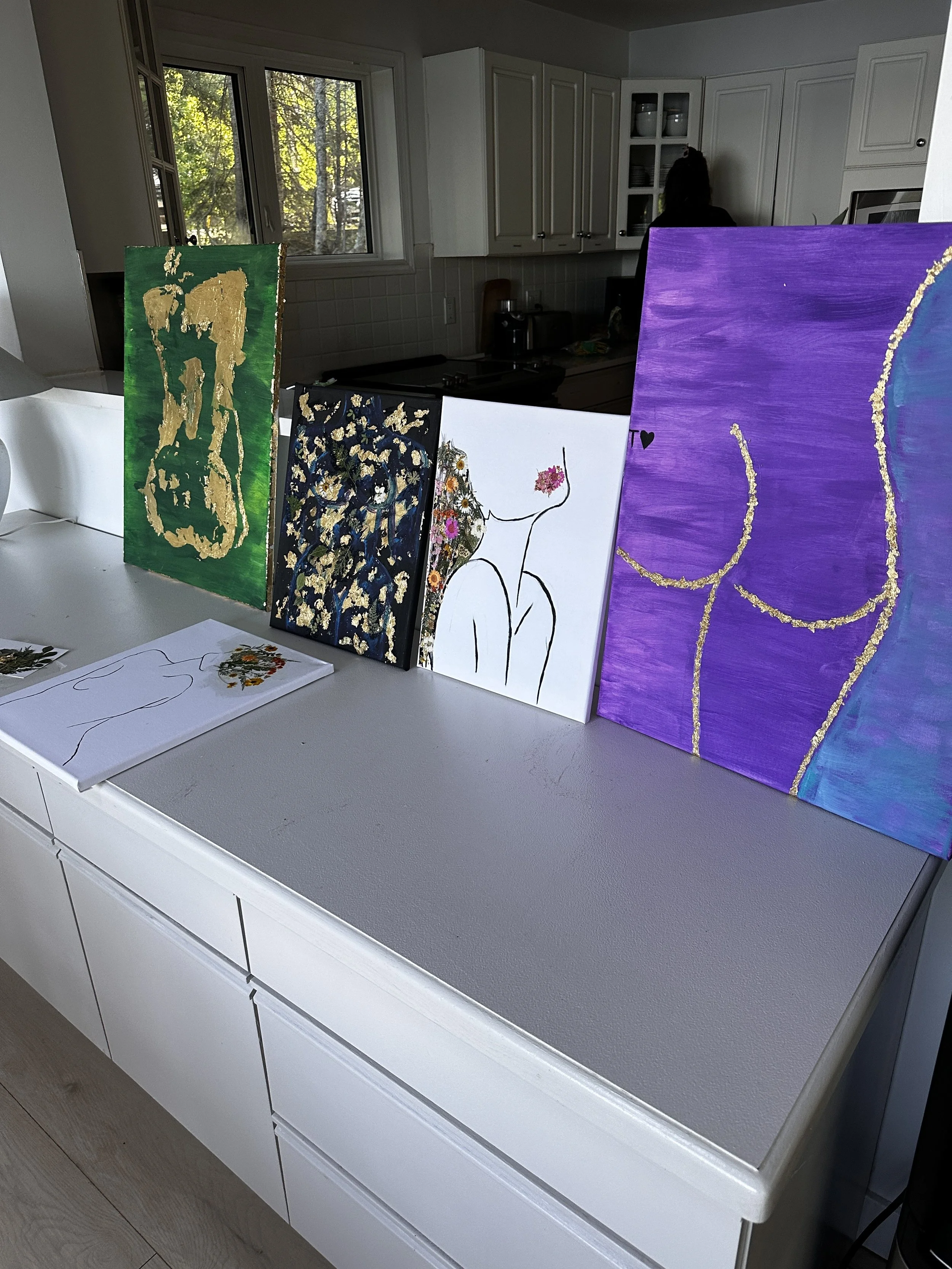 A series of four abstract paintings displayed on a kitchen countertop. The paintings feature colorful and textured designs, including gold accents, vivid colors like green, purple, and blue, and black outlines of human figures. The countertop is whit