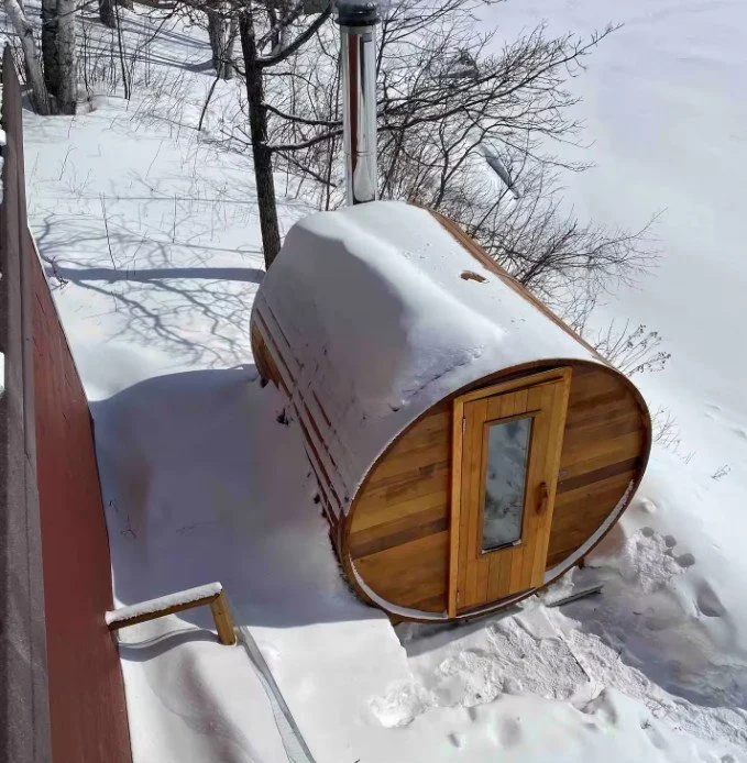 Wooden barrel sauna covered in snow next to a red building in a snowy landscape with trees.
