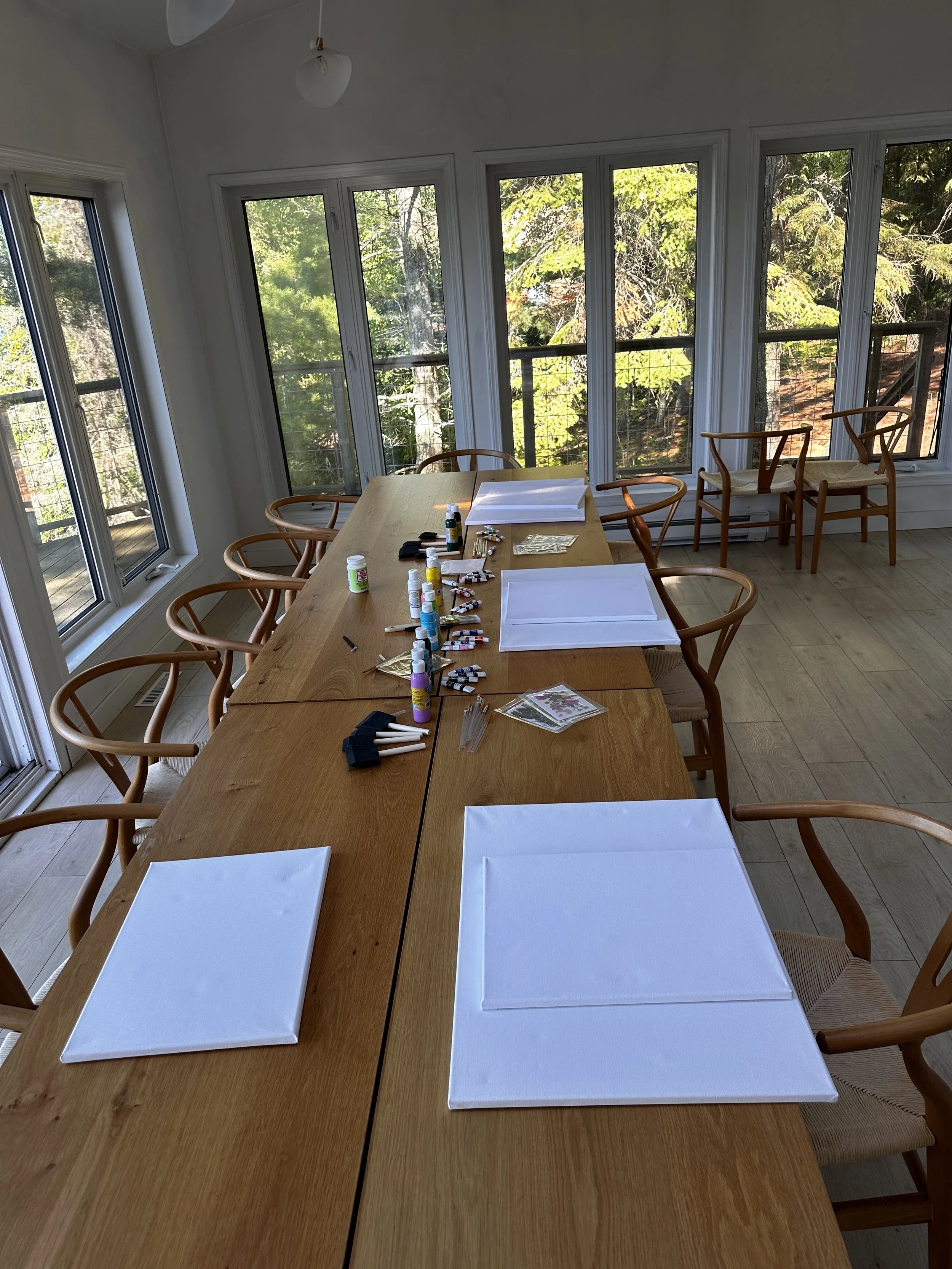 Room with a large wooden table set up for painting, featuring blank canvases, paintbrushes, paints, and other art supplies. Surrounded by windows with a view of trees outside.