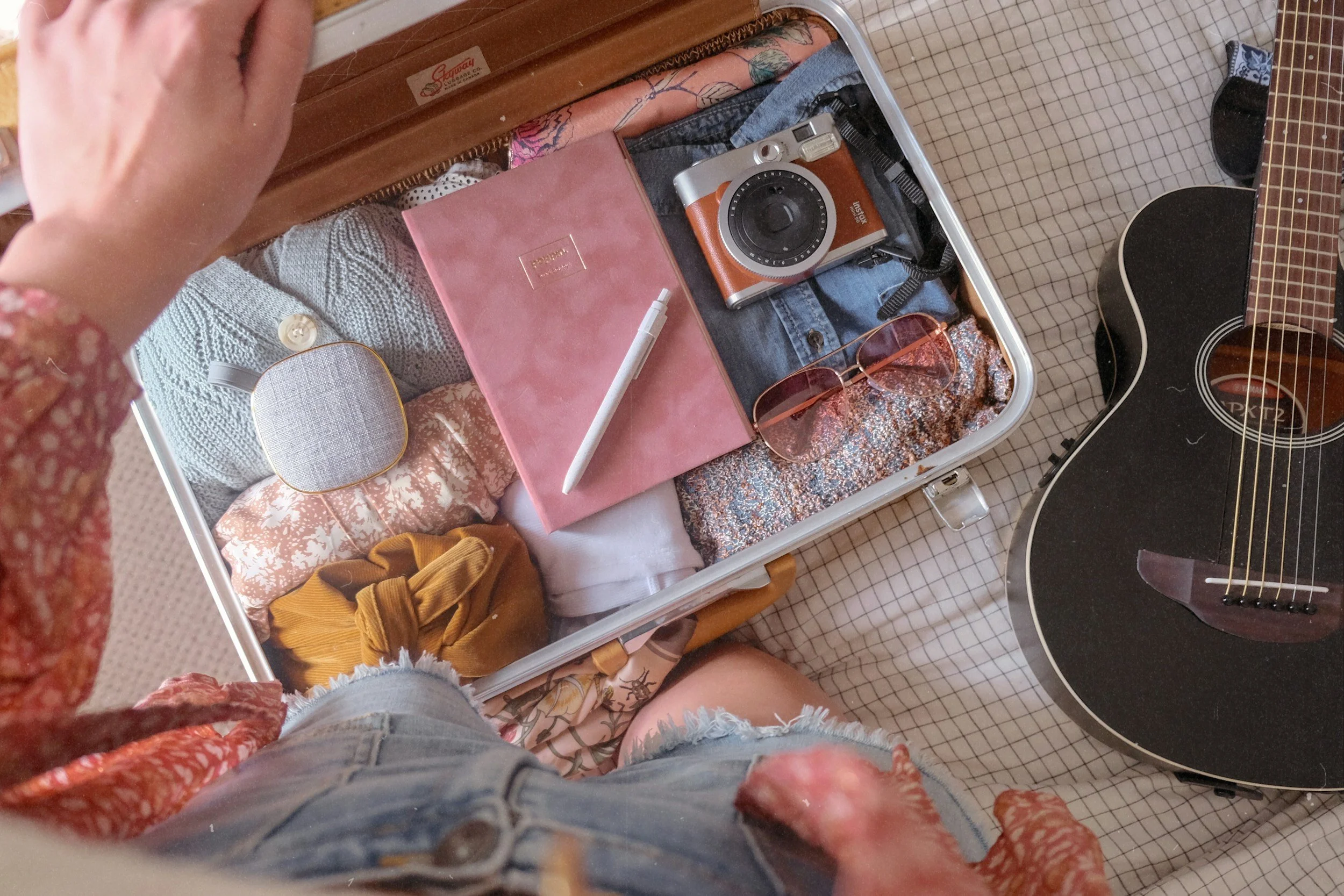 woman's open suitcase that she's packed that includes items like a vintage-look instant camera, journal and clothing