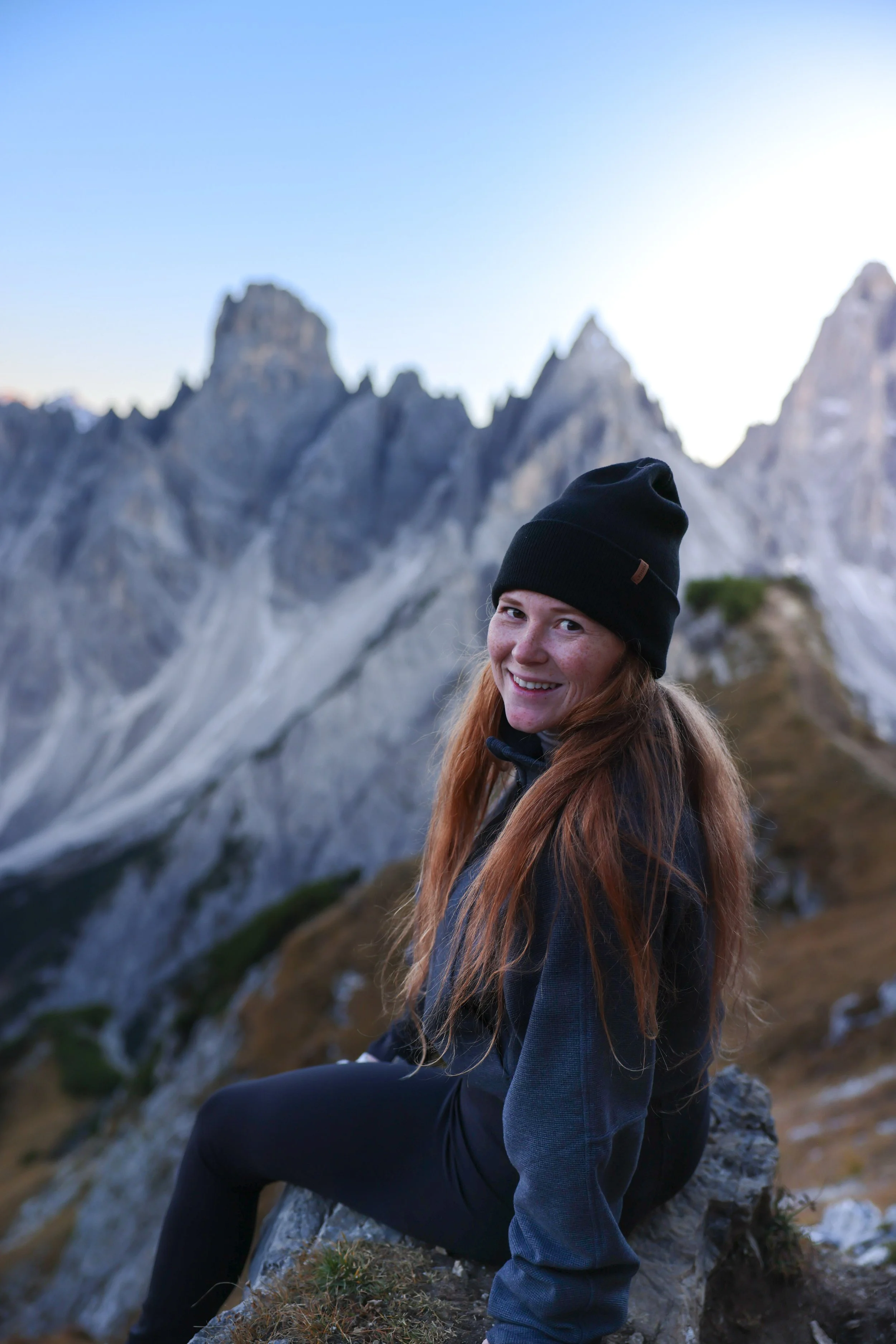 A smiling woman with long red hair, wearing a black beanie and outdoor jacket, sitting on a rock with mountain peaks in the background during daylight.