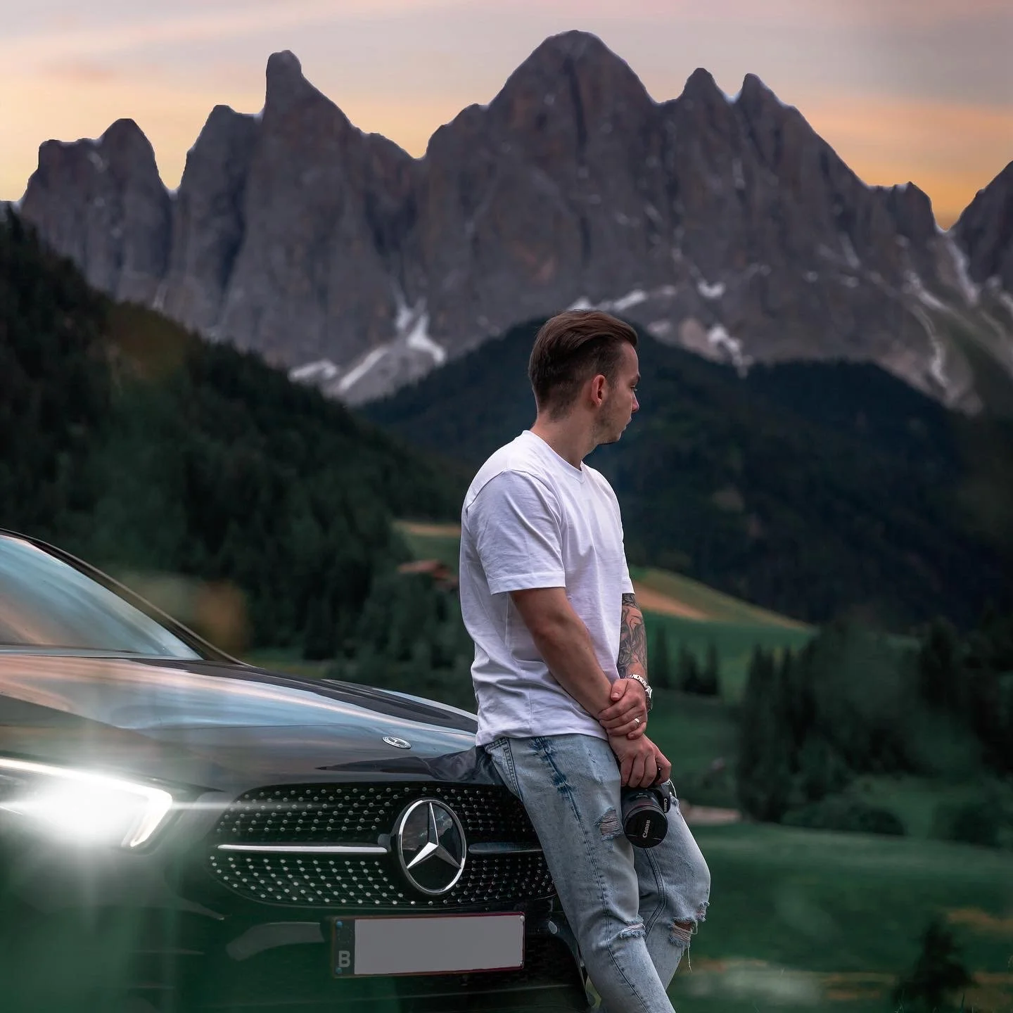 A young man in a white t-shirt and ripped jeans leaning against a black Mercedes-Benz car, holding a camera, with mountains and a sunset sky in the background.