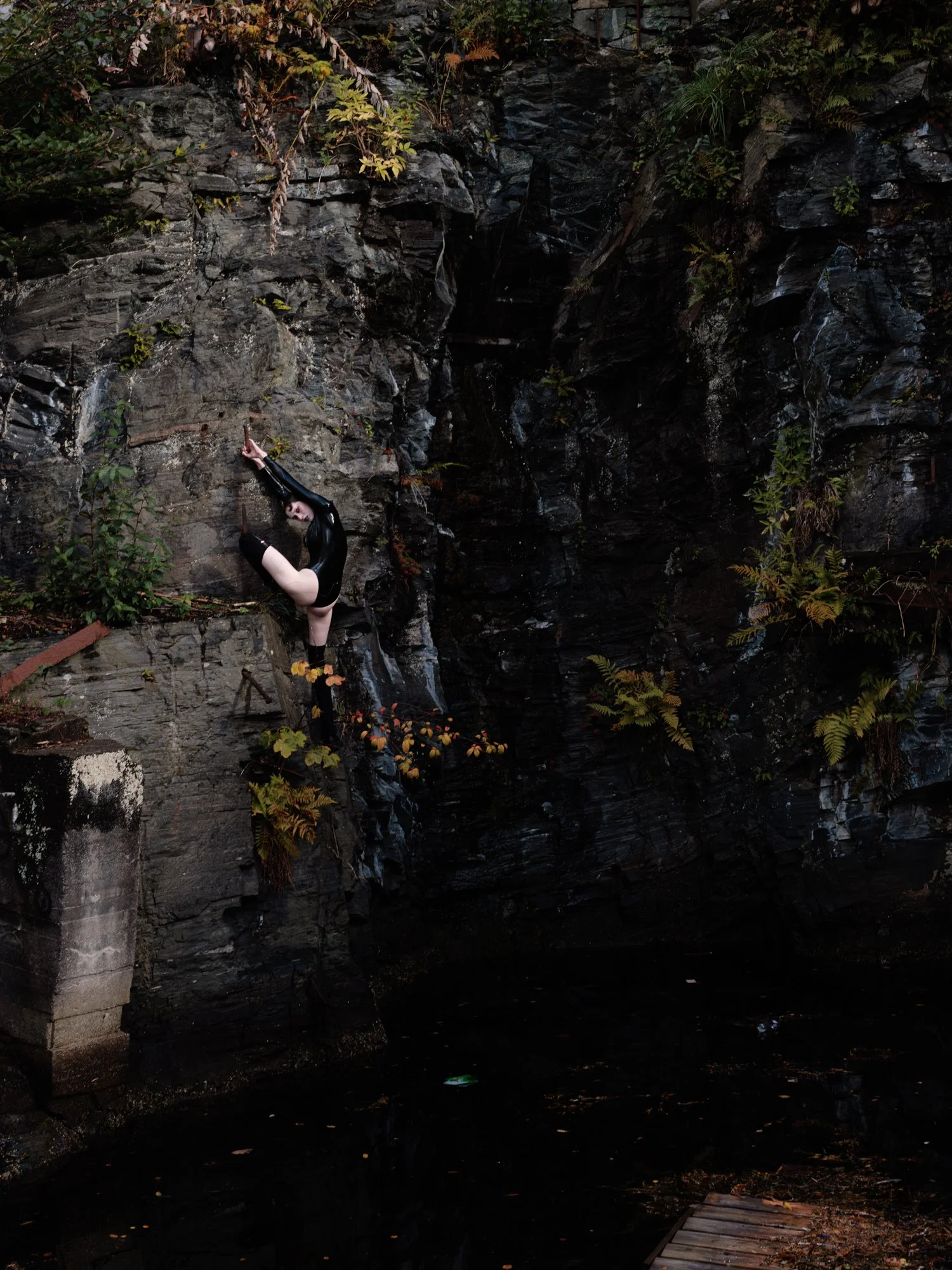 A woman in a black dress and long gloves performing a pose against a rocky cliffside near water, surrounded by greenery.