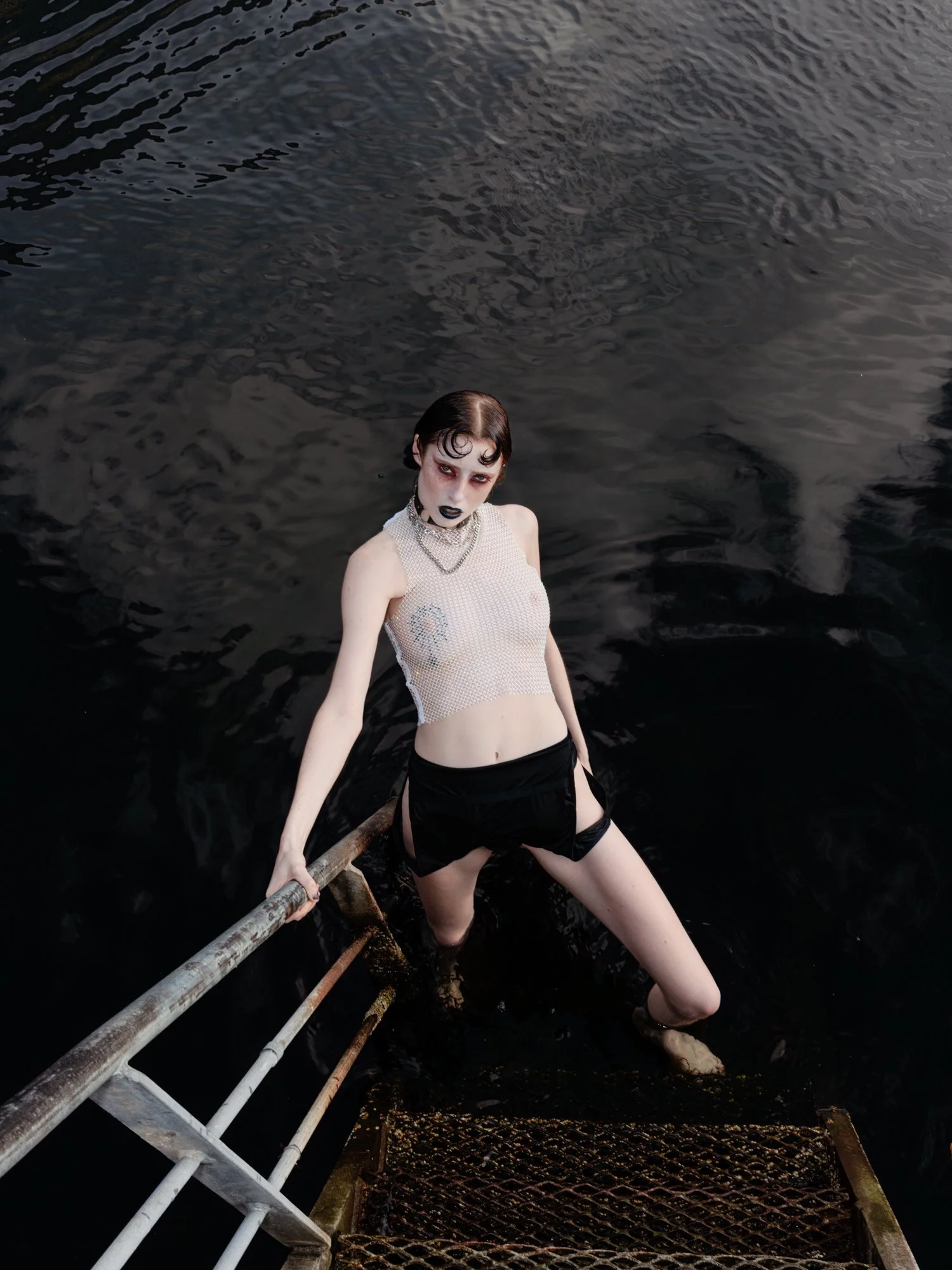 A woman with dark makeup and wet hair poses on a metal ladder extending into a dark body of water, wearing a mesh top, black shorts, and jewelry.
