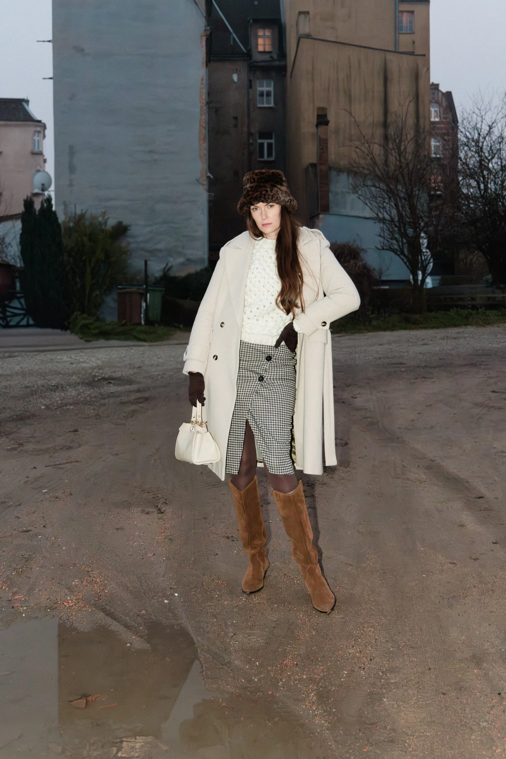 A woman standing outdoors on a dirt path, dressed in a cream coat, plaid skirt, brown knee-high boots, and a leopard print hat, holding a small white handbag.