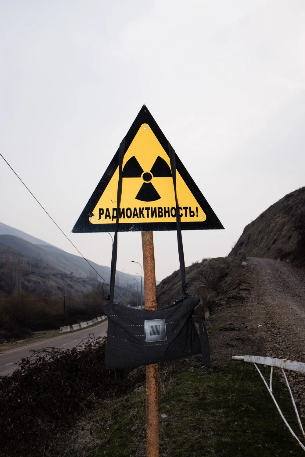 Yellow warning sign with a radiation symbol and Russian text, attached to a pole with a black bag hanging from it, set outdoors next to a dirt road and hillside.