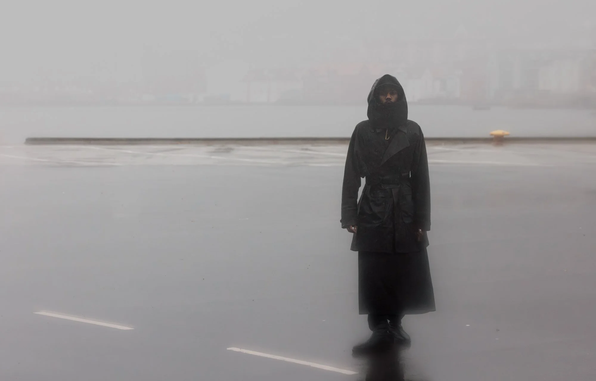 Person wearing a long black coat and headscarf standing alone on a foggy, empty street with overcast sky.