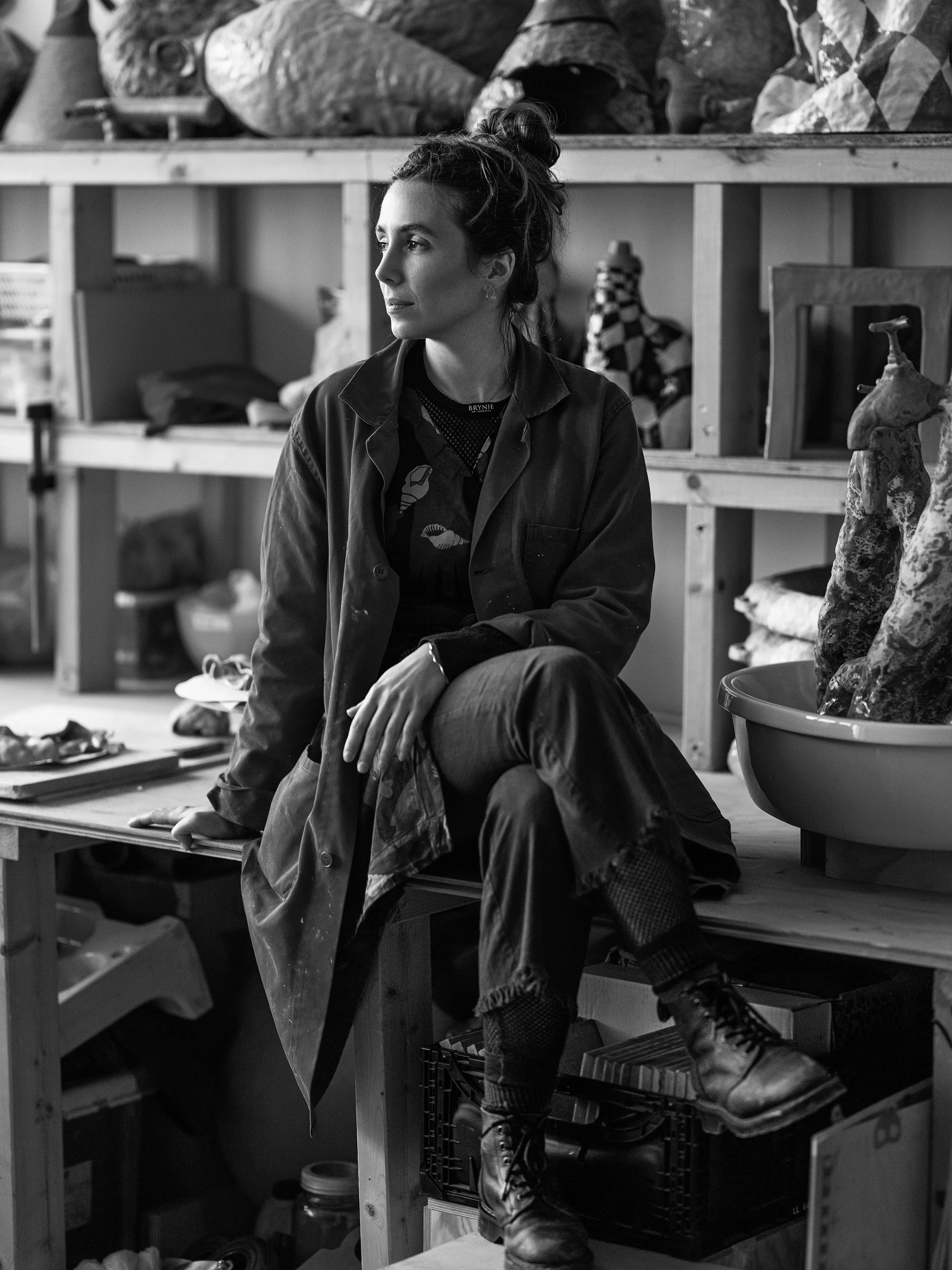 A woman with her hair in a bun sitting on a workbench in a cluttered room, looking to the left.