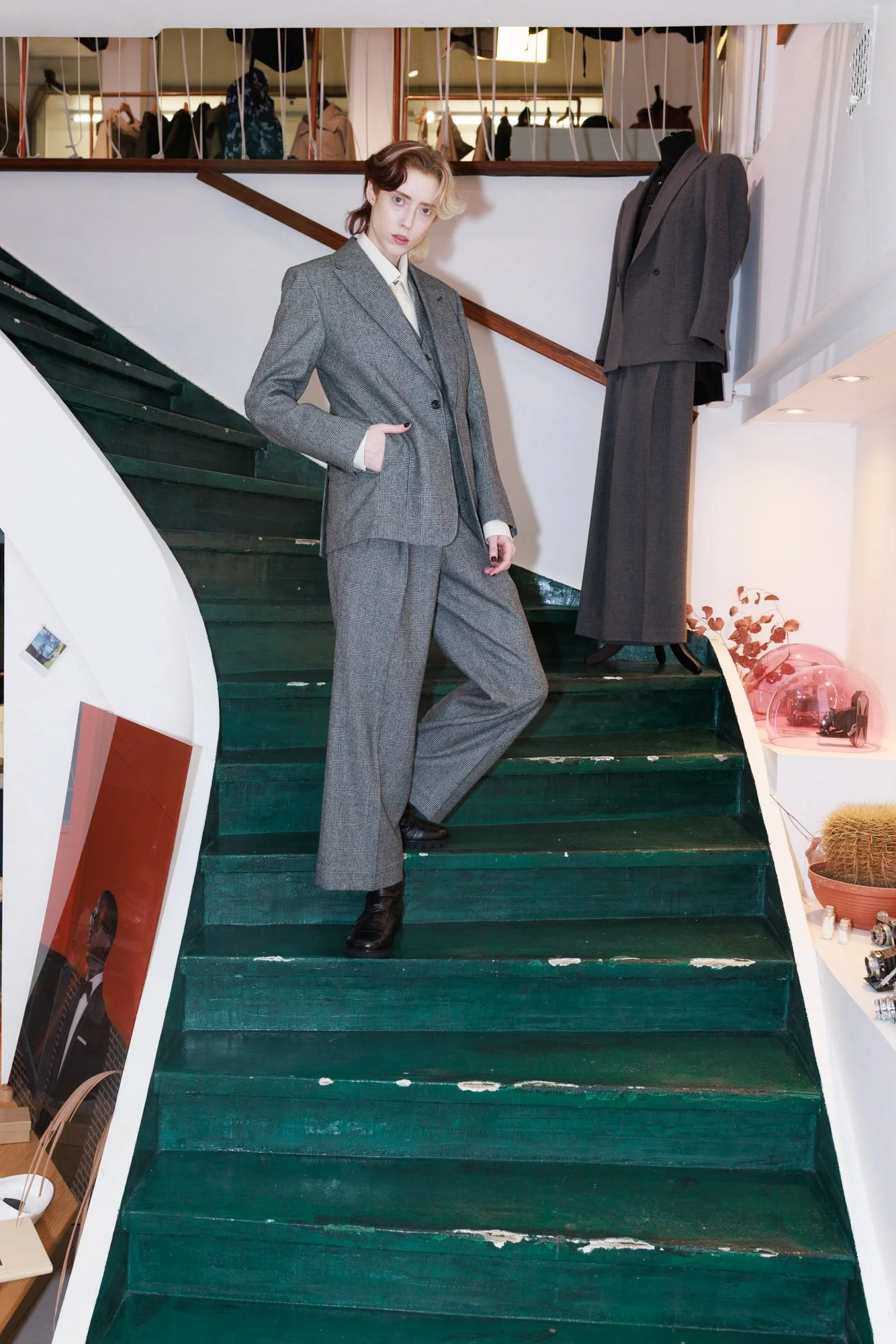 A woman in a gray plaid suit standing on a green staircase inside a clothing store, with another suit displayed on a mannequin behind her.
