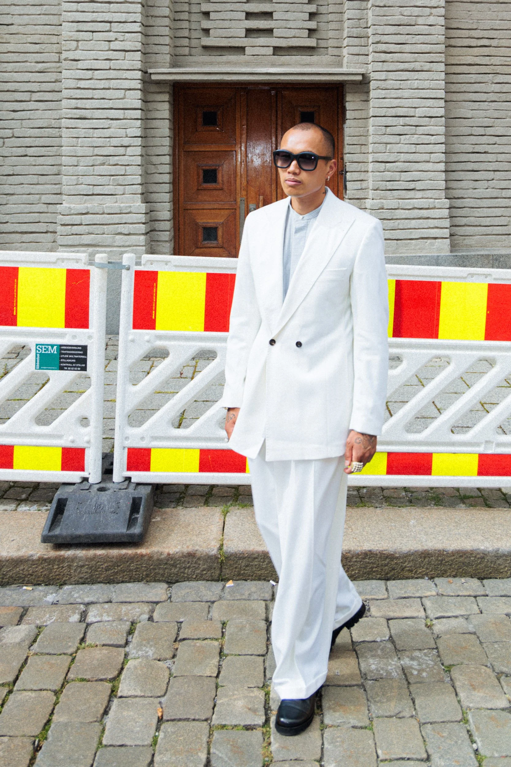 Person standing outdoors in front of a construction barrier and a brick building, wearing a white suit, sunglasses, and black shoes.