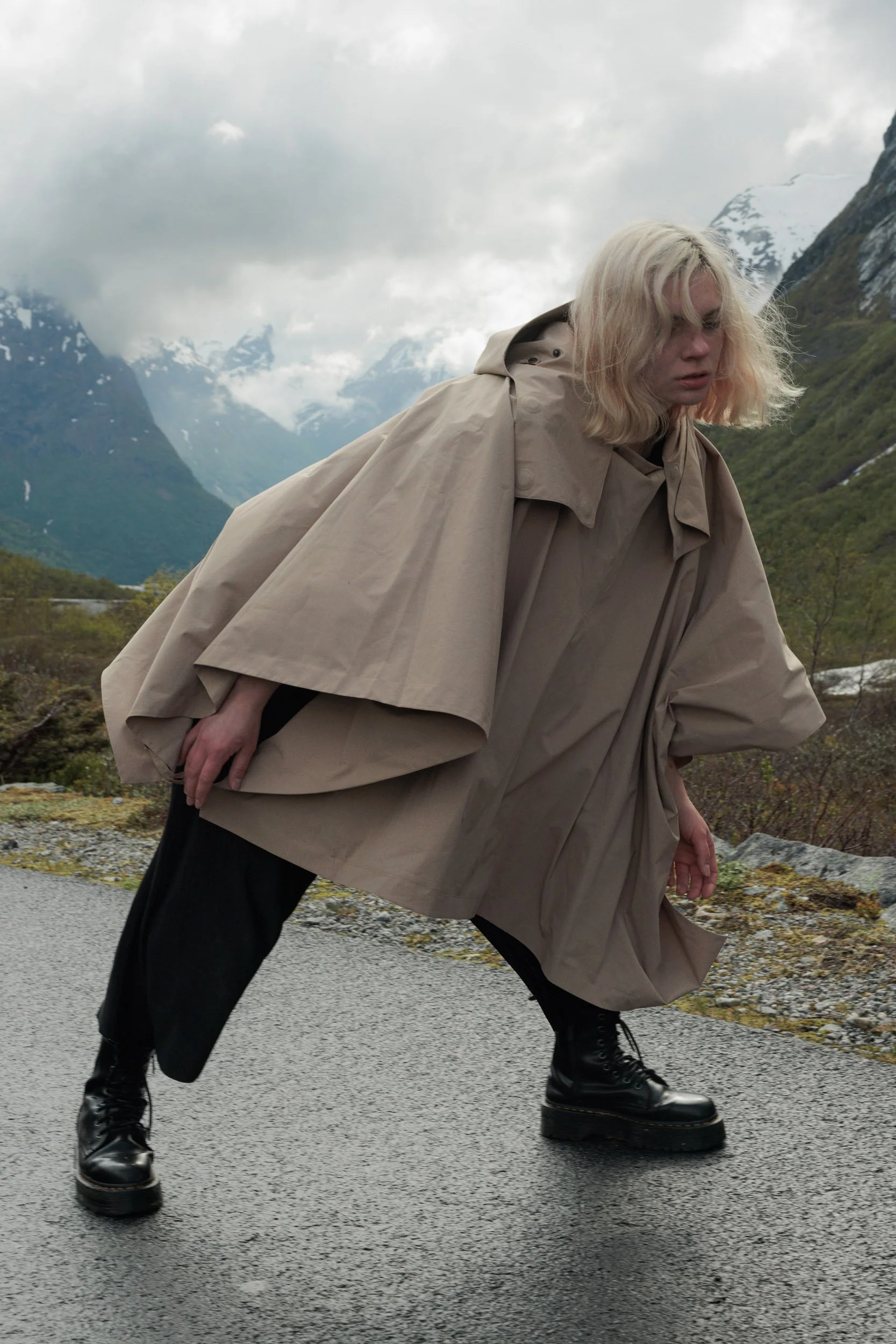 A woman wearing a beige oversized coat and black boots standing on a rainy mountain road with mountains and cloudy sky in the background.