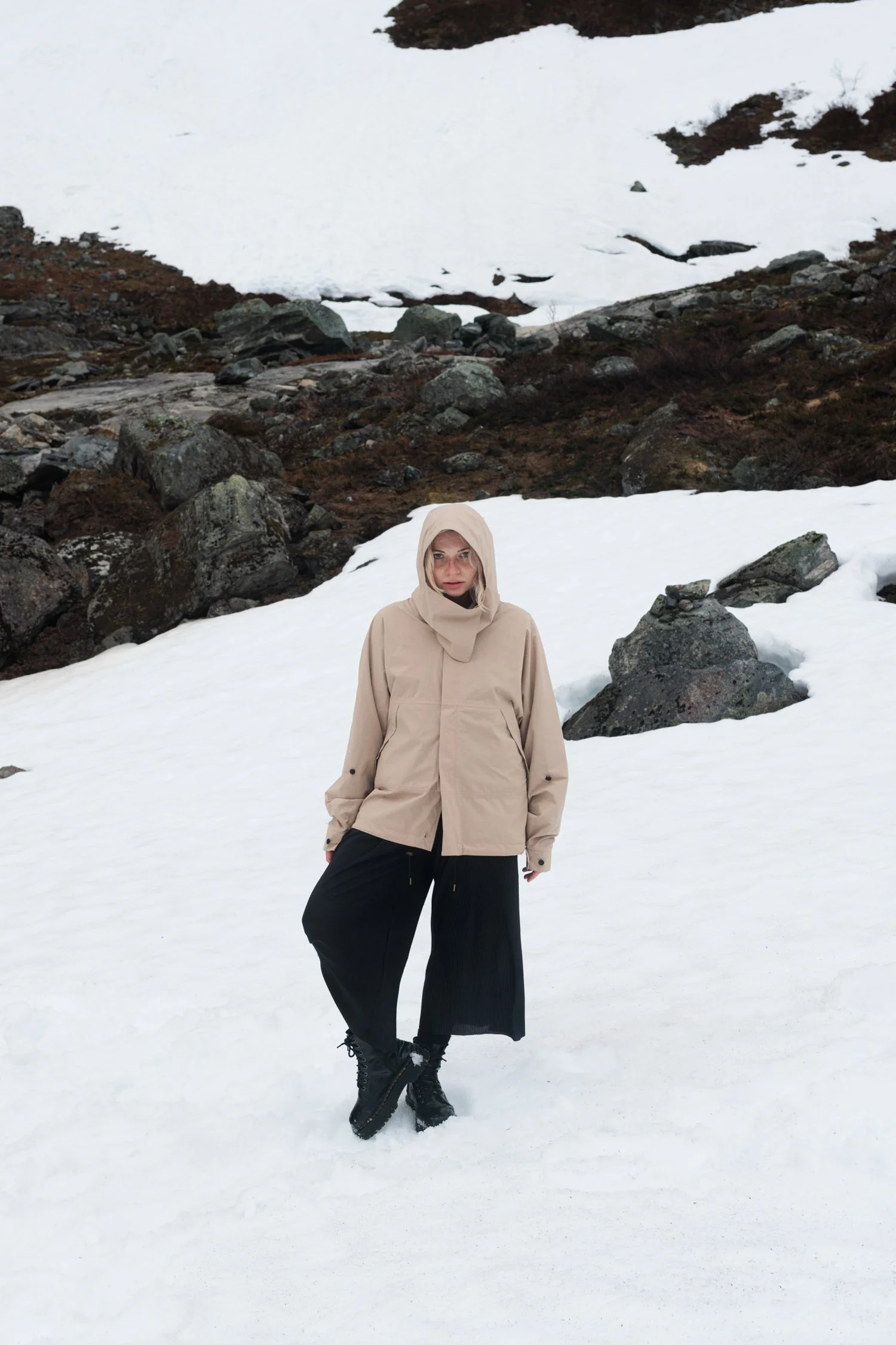 A woman standing in snow on a hillside with rocks and snow patches in the background, wearing a beige hooded jacket, black wide-leg pants, and black boots.