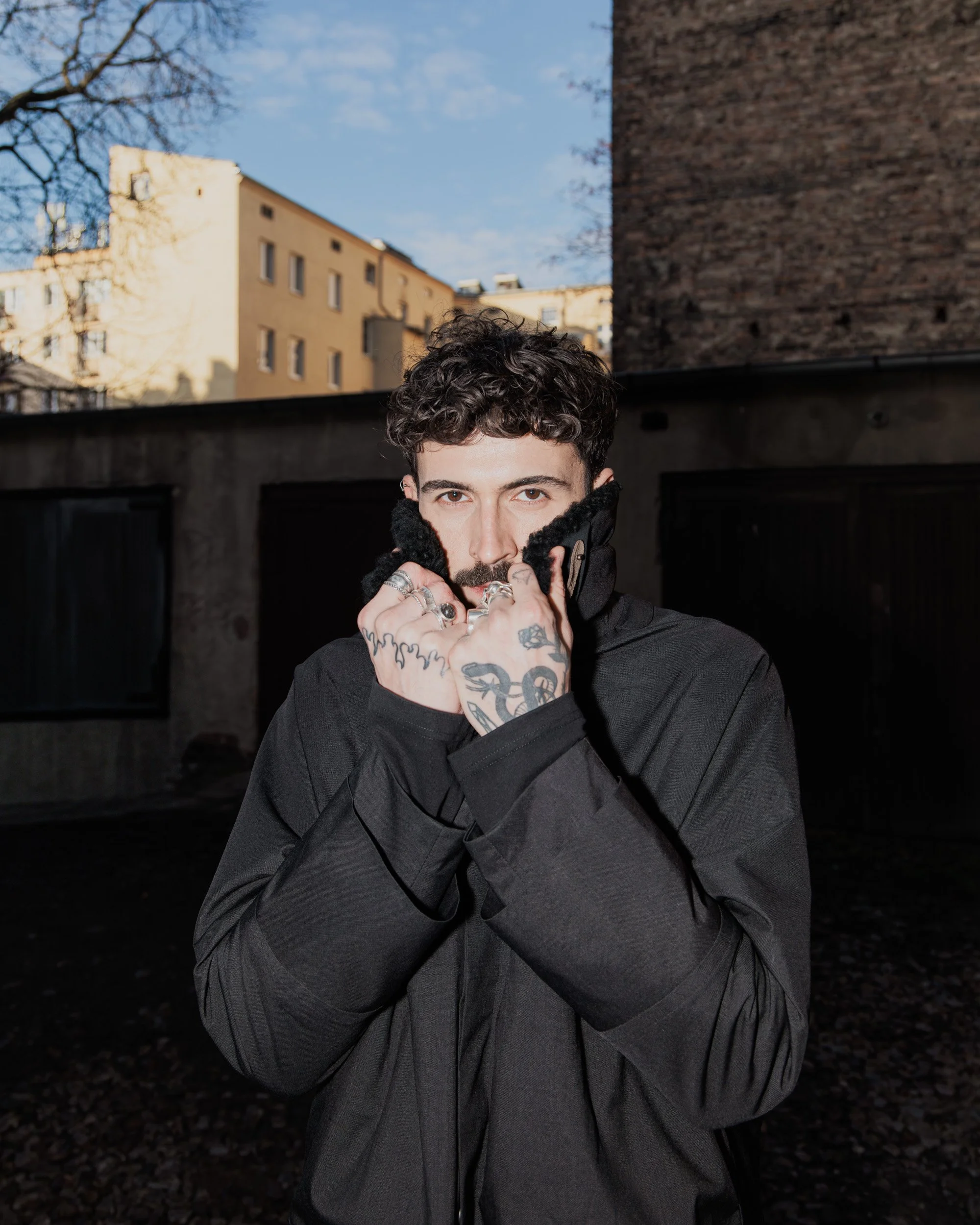 A man with curly dark hair and tattoos on his hands holding up the collar of his black jacket outdoors, with buildings and a blue sky in the background.