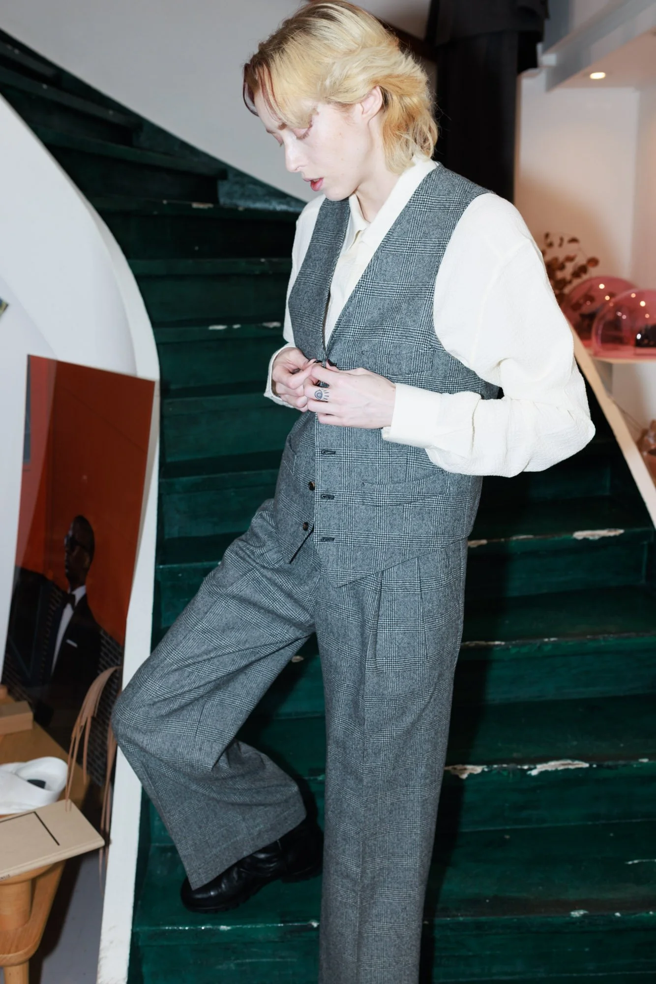 A person with blonde hair, dressed in a white shirt and gray plaid vest with matching wide-leg pants, standing on a dark green staircase indoors, looking down and adjusting their buttoned vest.