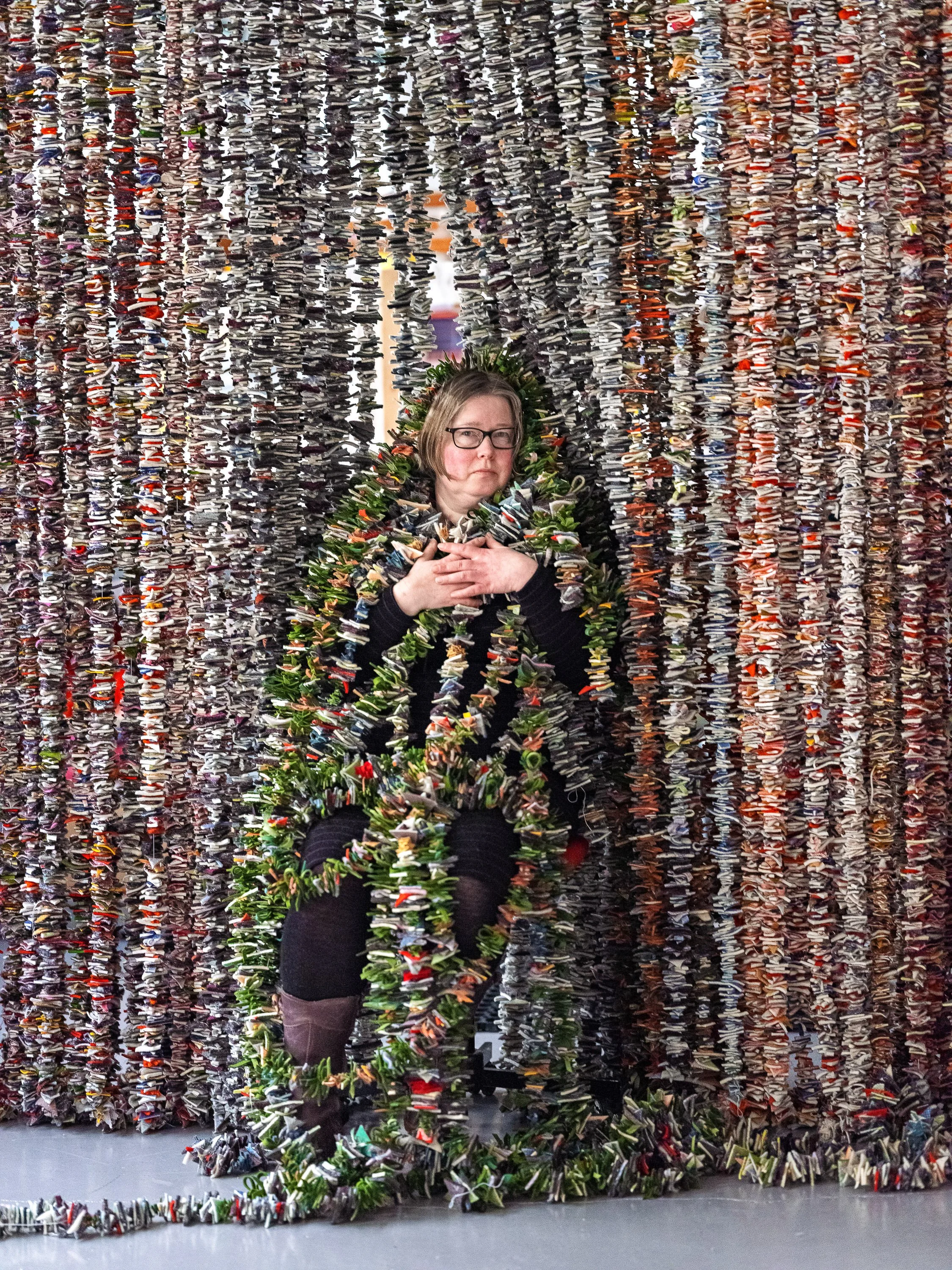 A woman with glasses posing amidst a large installation made of numerous paper shoes, with her arms crossed and a neutral expression.