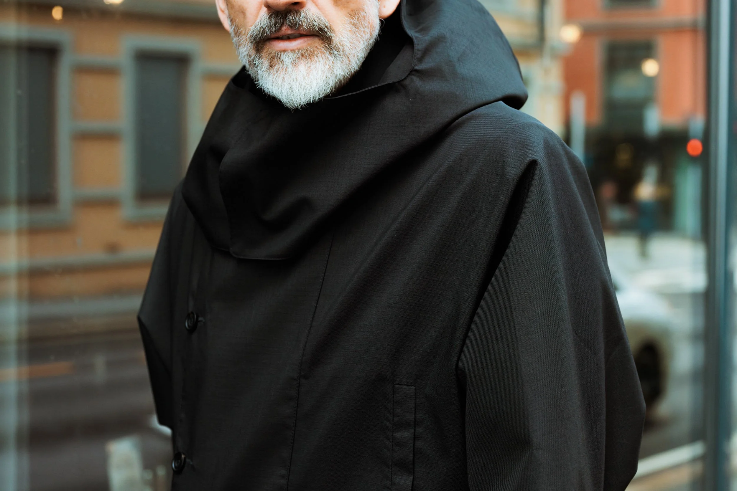 Close-up of a man with a gray beard wearing a black coat and scarf, standing in front of large windows with city buildings visible in the background.