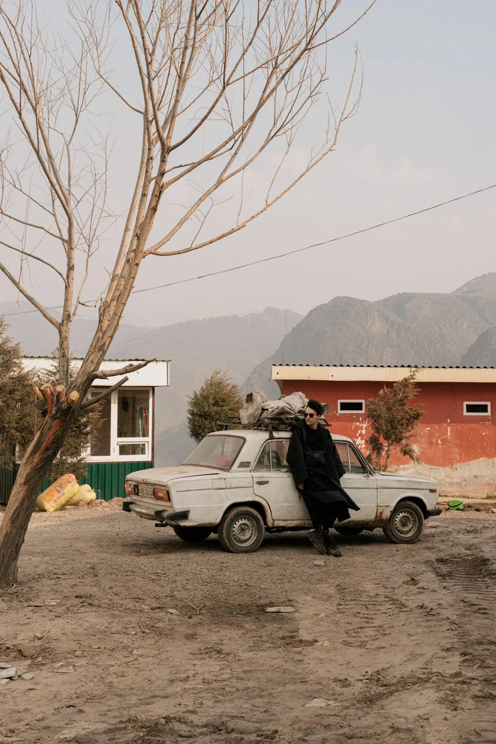 A person in black clothing and sunglasses leaning against an old, rusty car with luggage on top, in a rural area with mountains in the background, a leafless tree, and a red and green building.