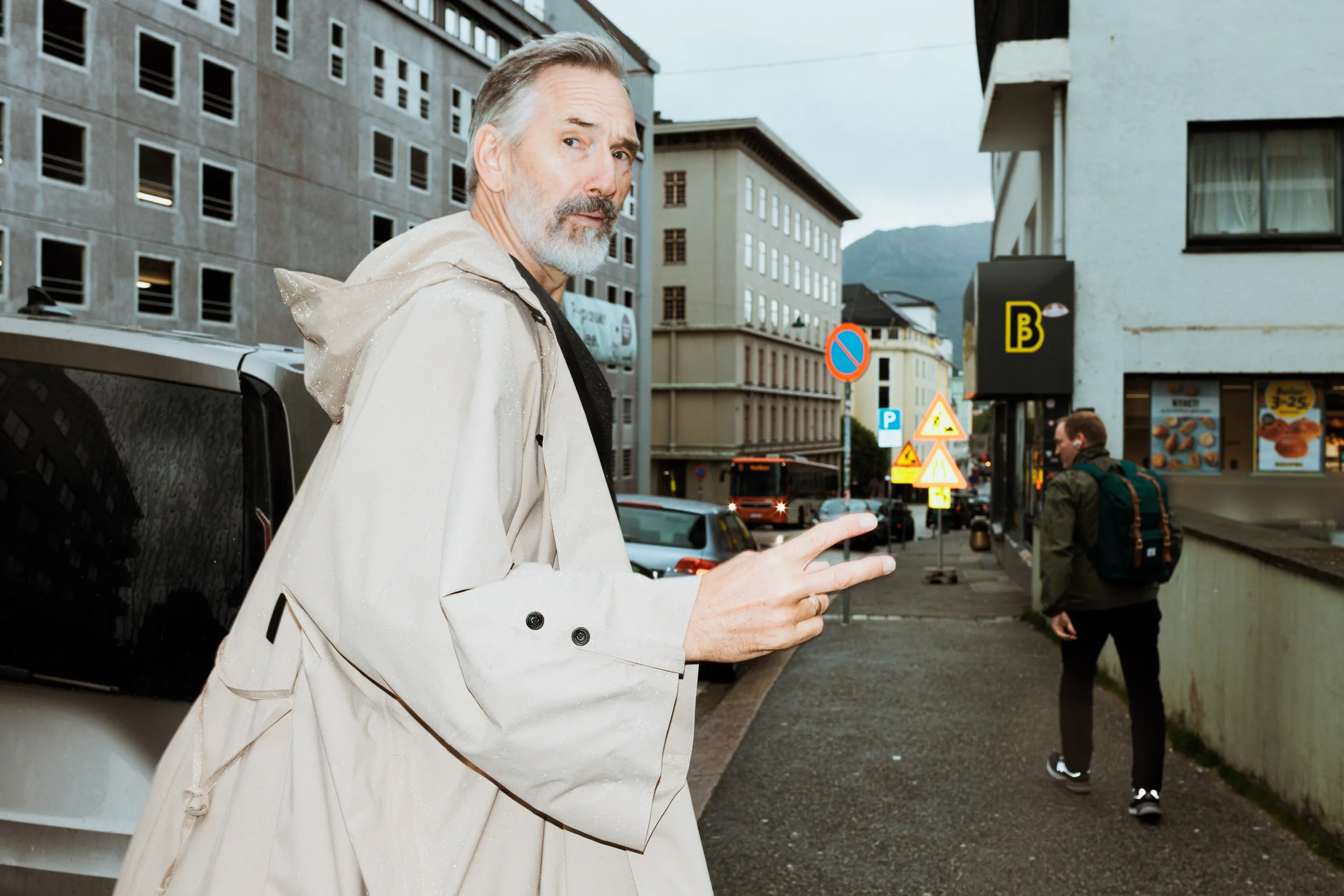 A man with gray hair and a beard making a peace sign with his fingers while standing on a city sidewalk during daytime, with a young man with a backpack walking away in the background and various traffic signs and buildings around.