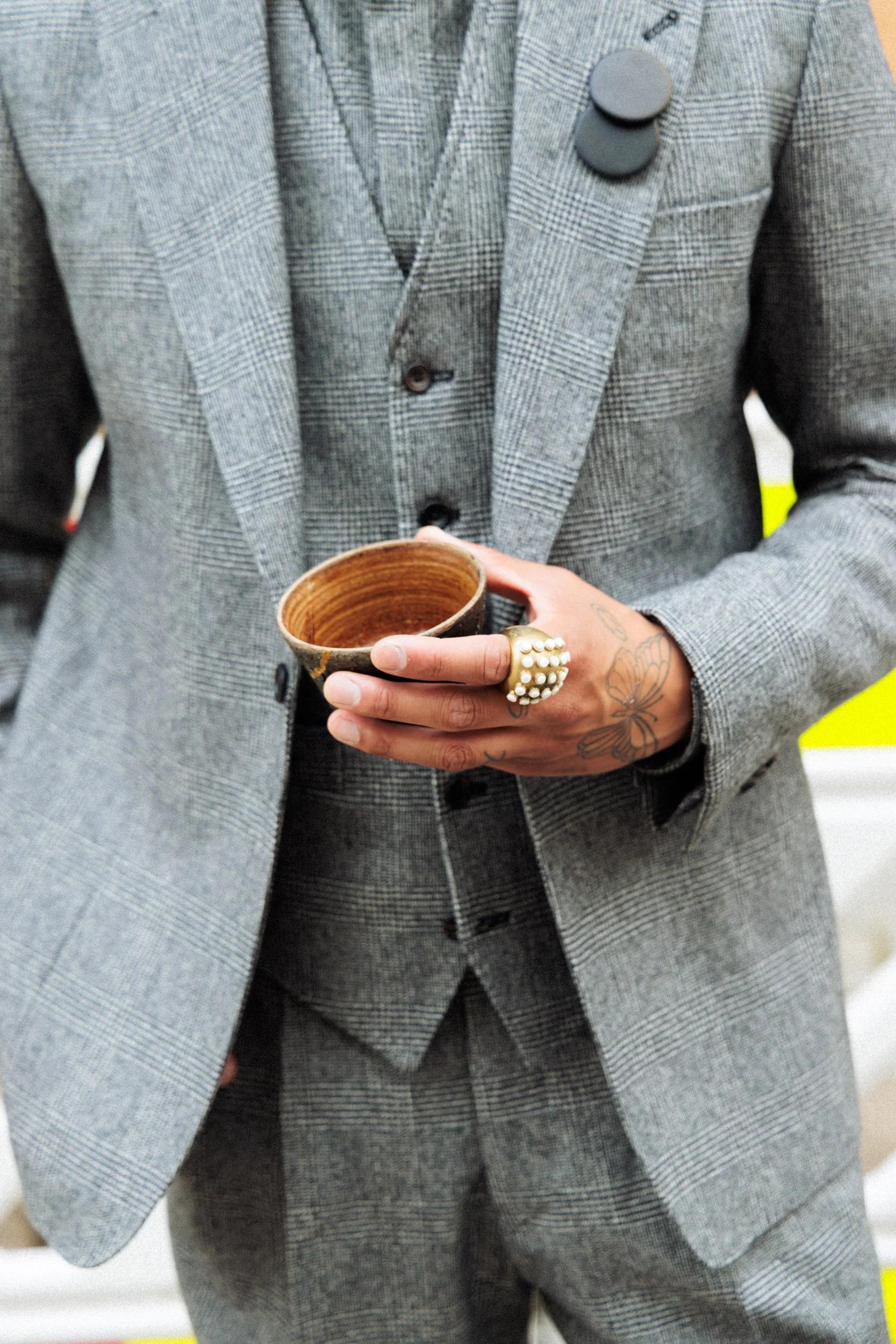 Person in a gray plaid suit holding a small ceramic bowl, wearing rings and tattoos on fingers and hand.
