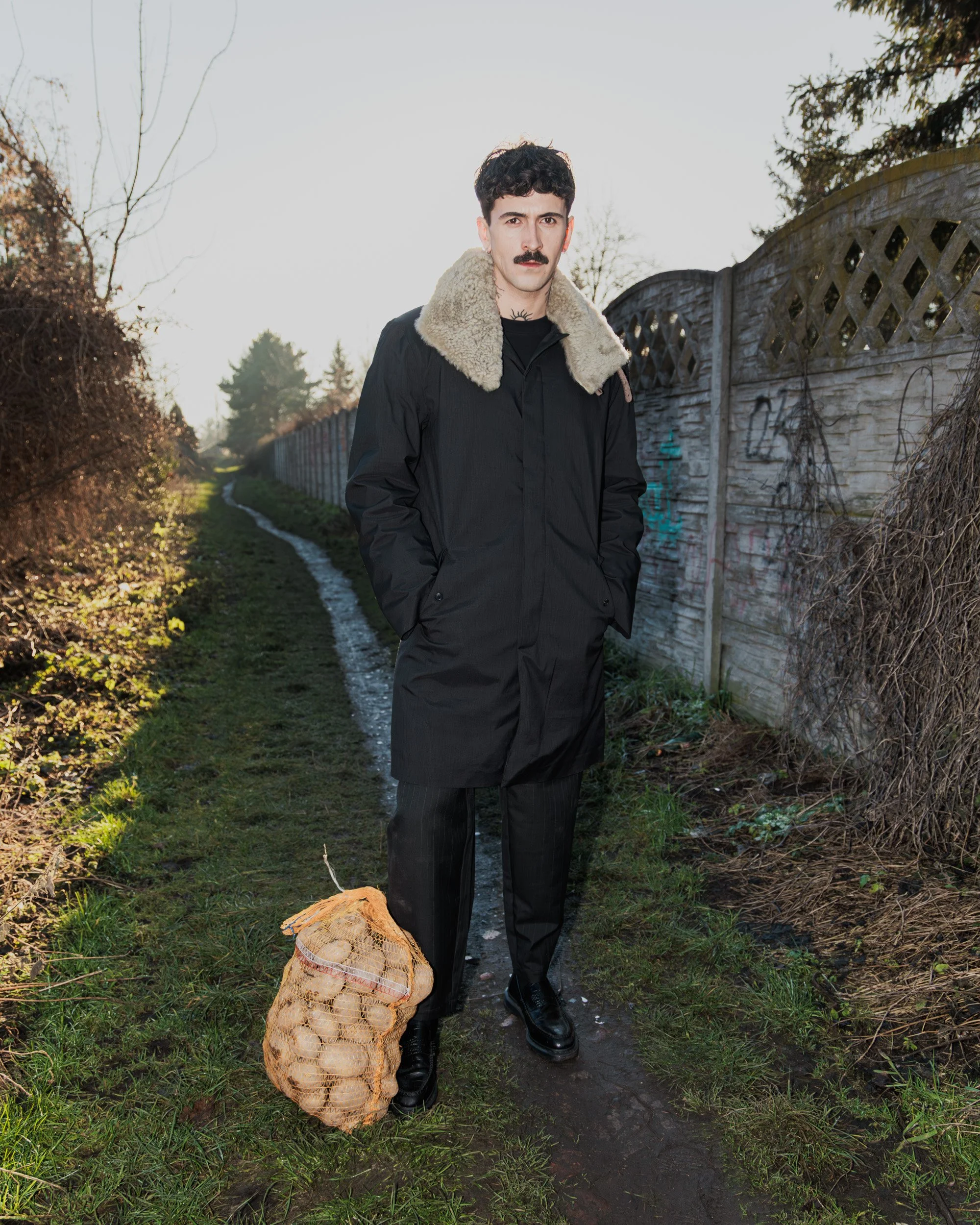 A man with dark hair, a mustache, and a serious expression, standing on a muddy path beside a wooden fence, holding a bag of potatoes.