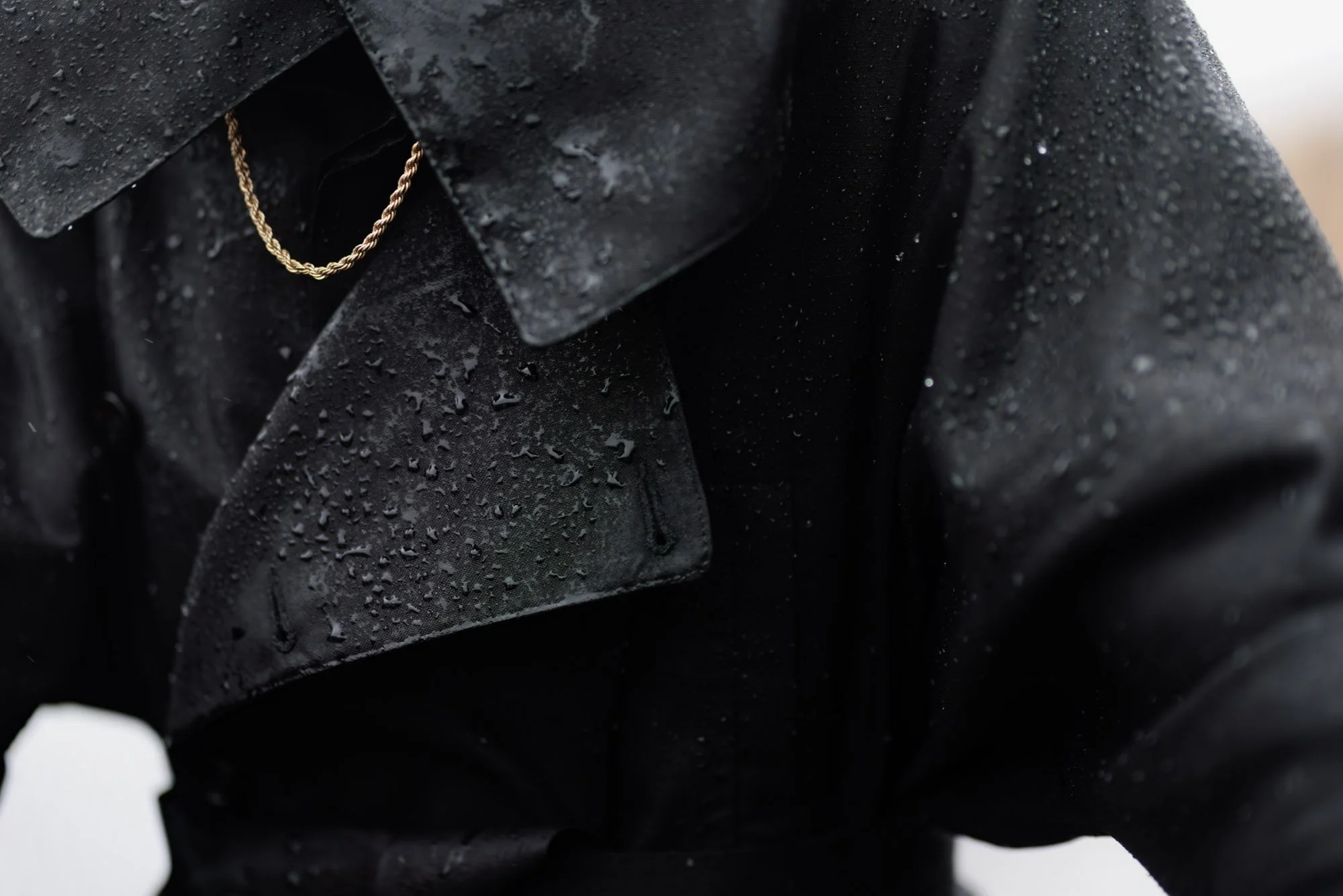 Close-up of a black raincoat with water droplets on its surface, featuring a leather collar and a gold chain necklace.