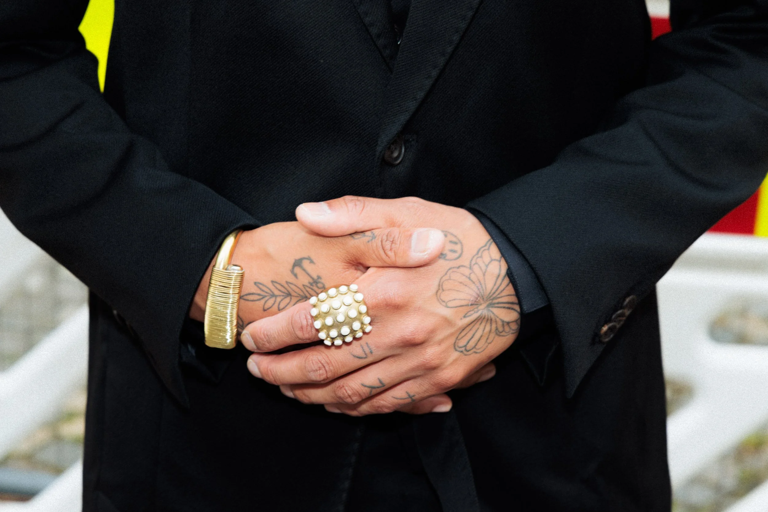 Person wearing a black suit with tattooed hands, gold bracelet, and a large pearl ring, clasping their hands together.