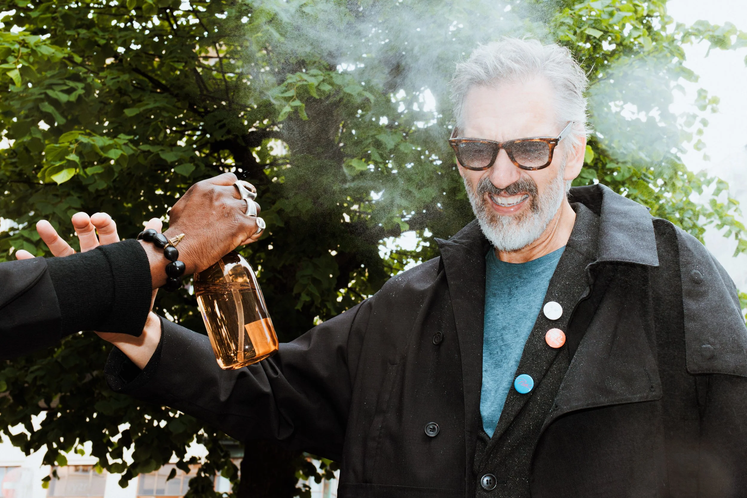 A man with gray hair and a beard, wearing sunglasses, a black jacket, and a blue shirt, smiling as another person hands him a glass bottle of beer outdoors with green trees in the background.