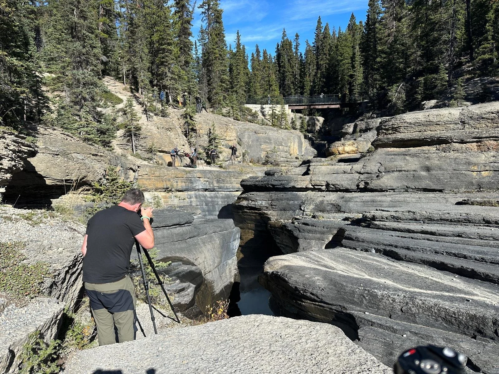 Man taking a photograph with a tripod beside a rocky canyon in a forested area, with multiple hikers on a trail above.