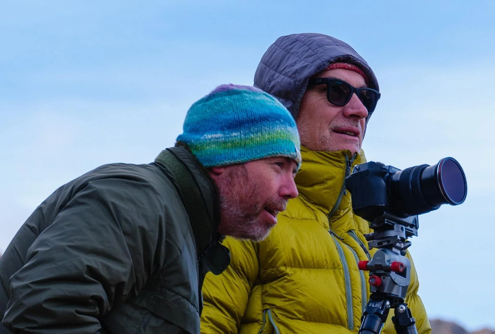 Two men dressed in winter jackets, one in a gray jacket with a multicolored beanie, and the other in a yellow jacket with sunglasses and a gray hood, looking through a telescope outdoors in cold weather.