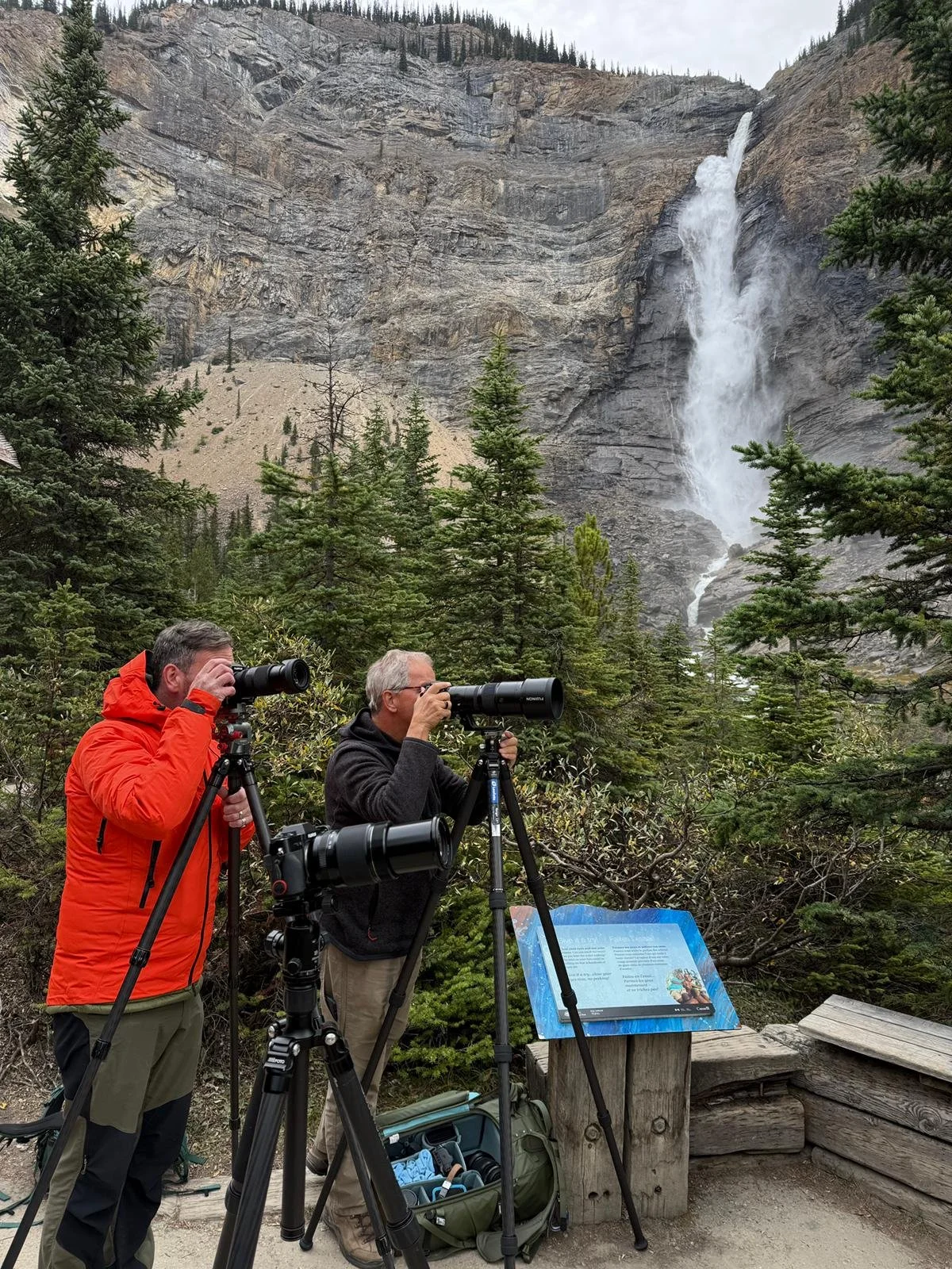 Two men with binoculars and telescopes observing a waterfall in a forested mountainous area, with informational signage nearby.