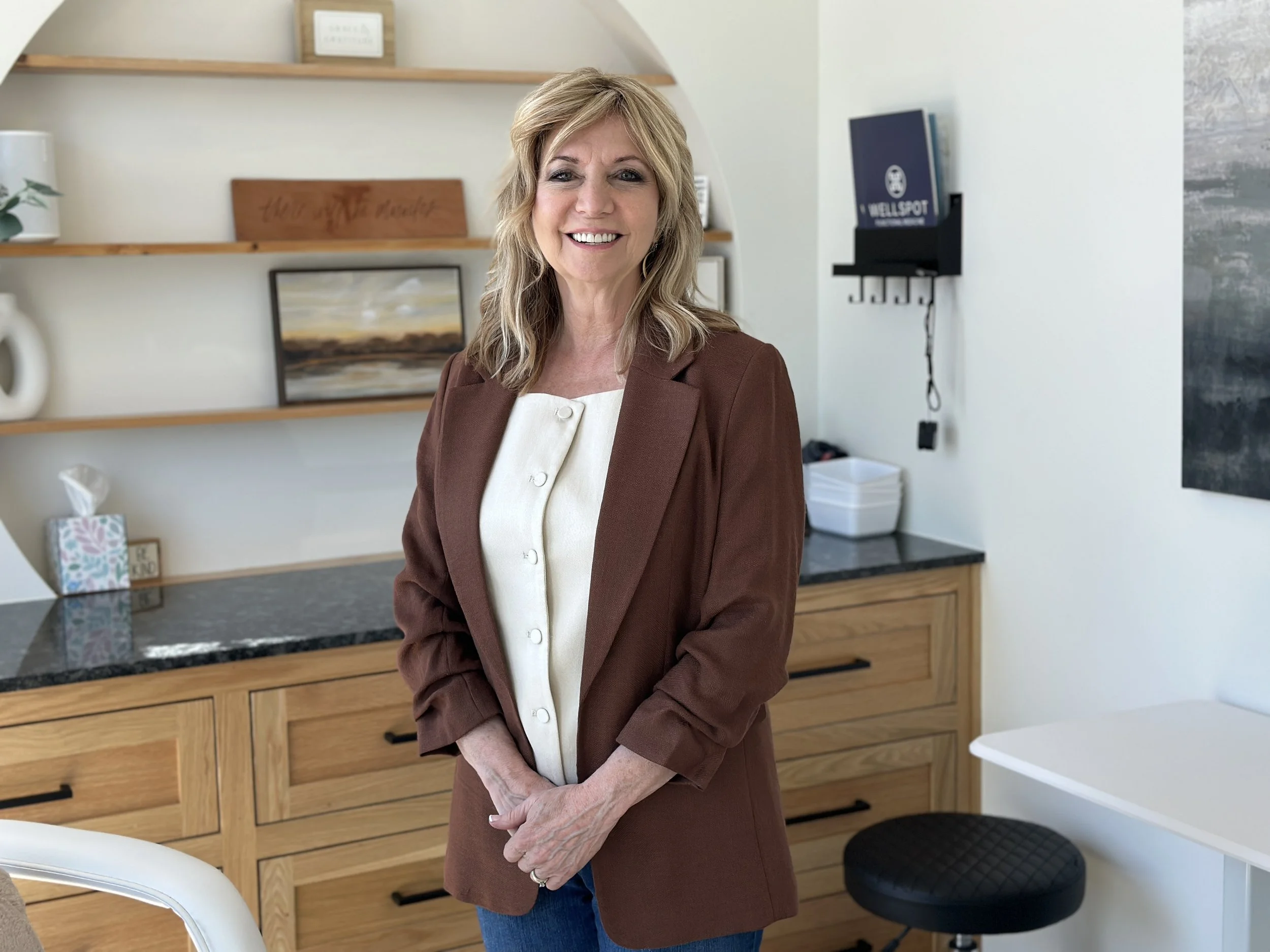 A woman with blonde hair dressed in a brown blazer and cream blouse, smiling in a modern kitchen with wooden cabinets, a bookcase, and a painted landscape on the wall.