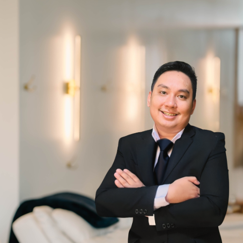 A man in a black suit and tie smiling with arms crossed in a well-lit indoor setting.