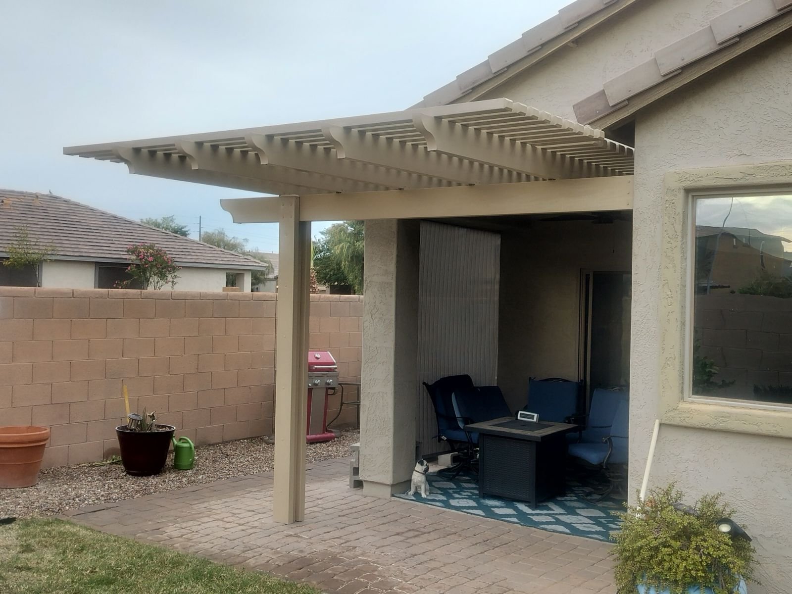 Backyard patio area with a covered seating space, blue chairs, and a dog sitting on a blue and white patterned rug. There is a small potted plant and a white pole near the seating area, and a barbecuer grill outside the fenced yard.