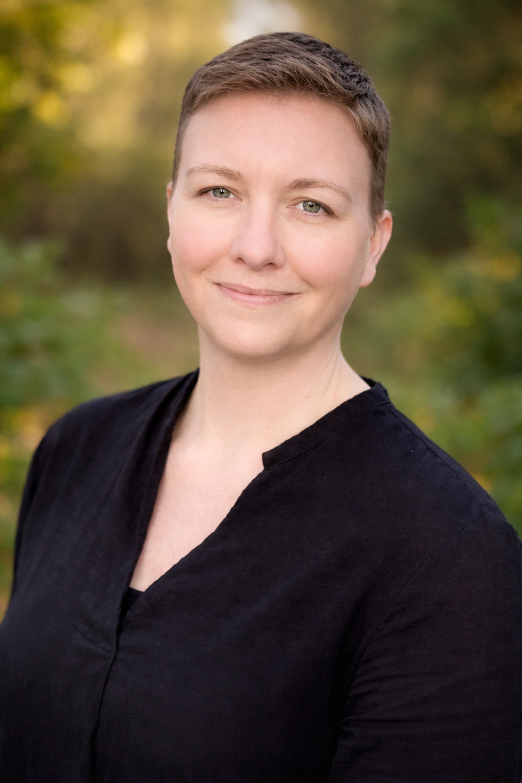 A woman with short brown hair and fair skin smiling outdoors, wearing a black top, with a blurred green and yellow natural background.