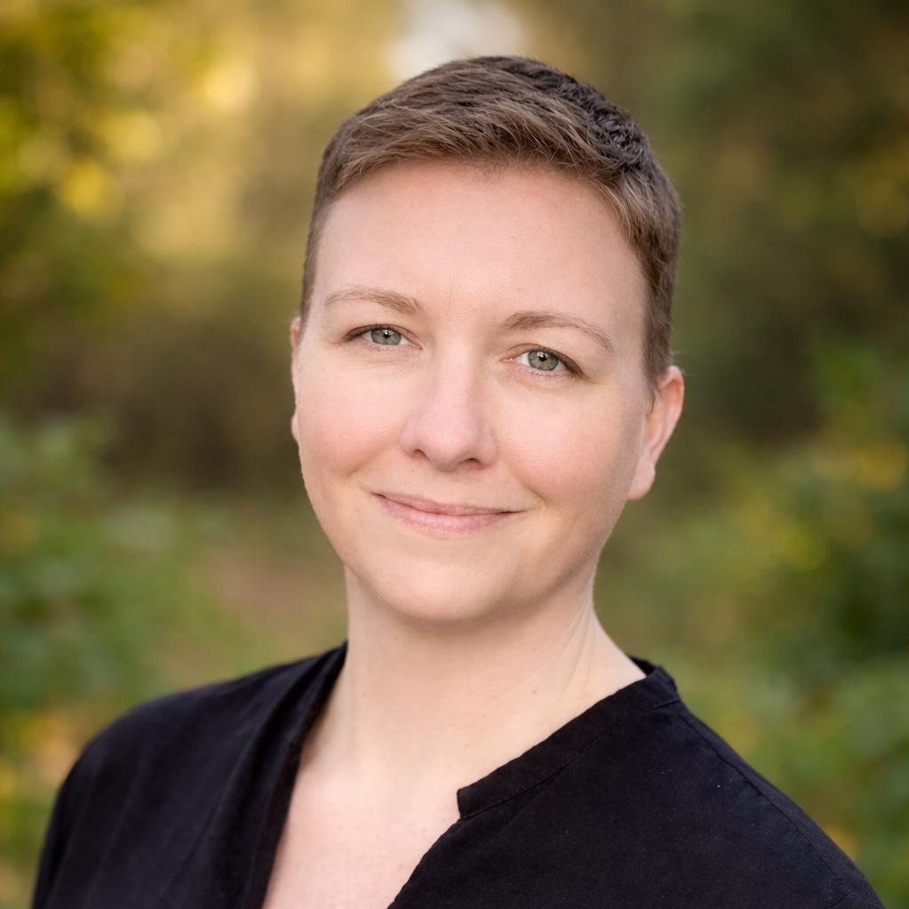 Portrait of a woman with short brown hair and light-colored eyes, smiling outdoors with a blurred background of green foliage.