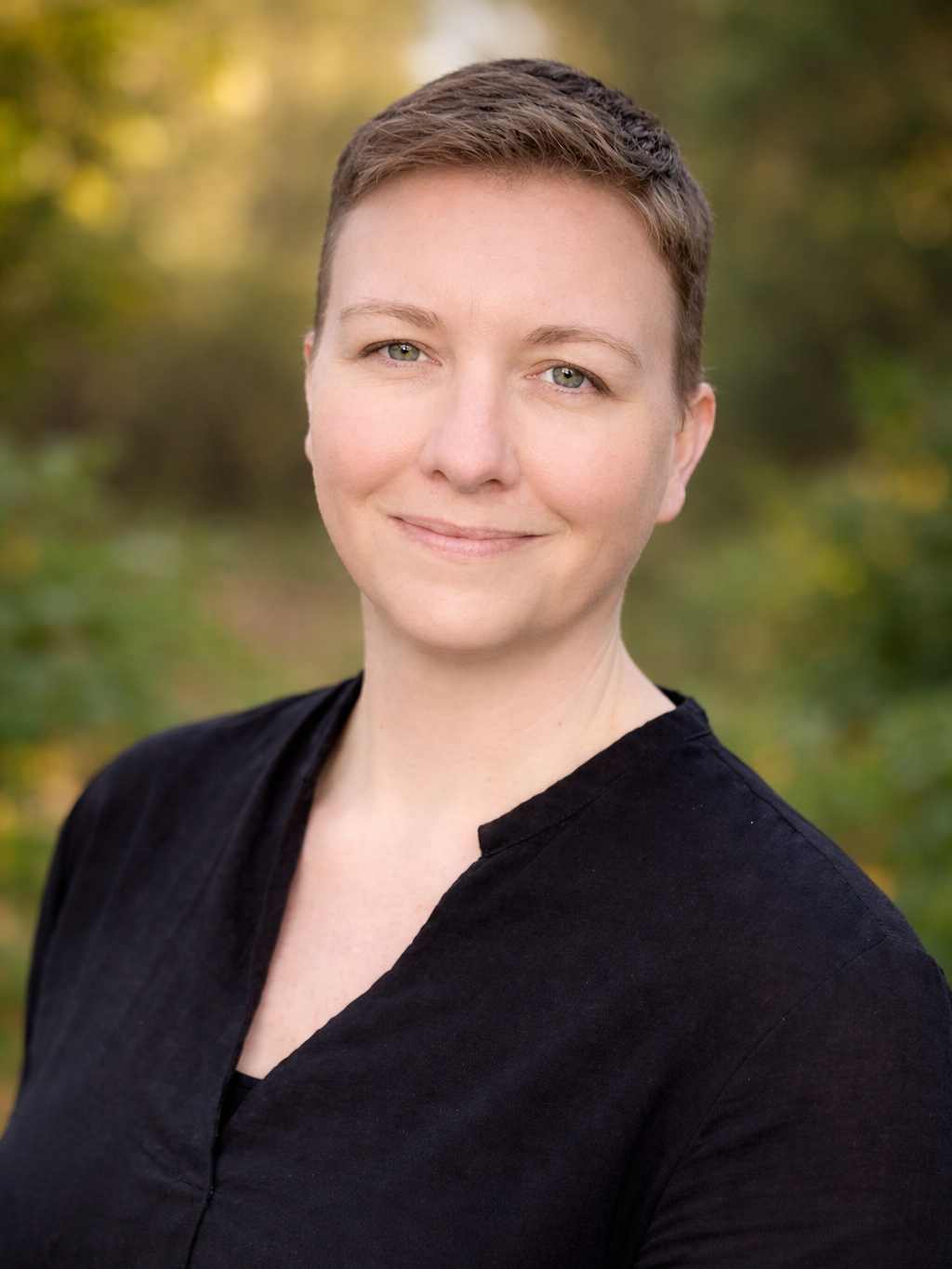 A woman with short brown hair, wearing a black top, smiling outdoors with blurred trees and greenery in the background.