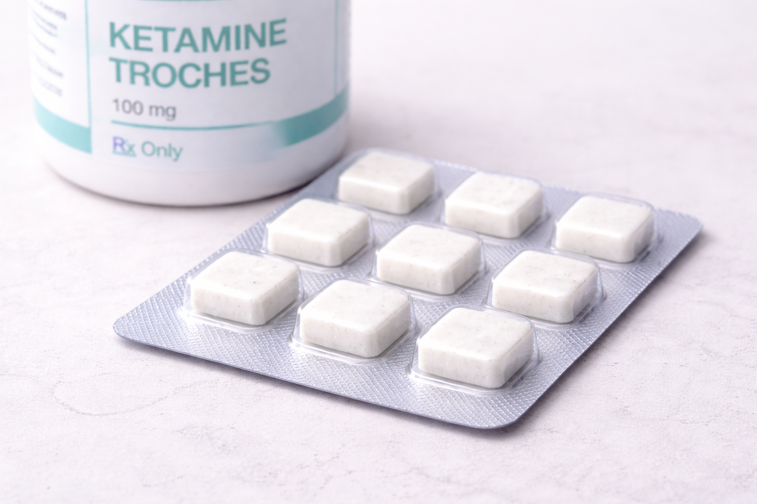 Blister pack of white ketamine troches in front of a white medicine bottle labeled 'Ketamime Troches 100 mg Rx Only'.