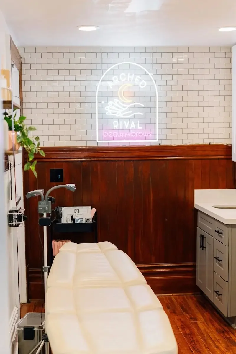 Treatment room at The State Co in Livermore, CA featuring a massage table, soft lighting, and a neon sign for Aesthetic Rival beauty and brow services.