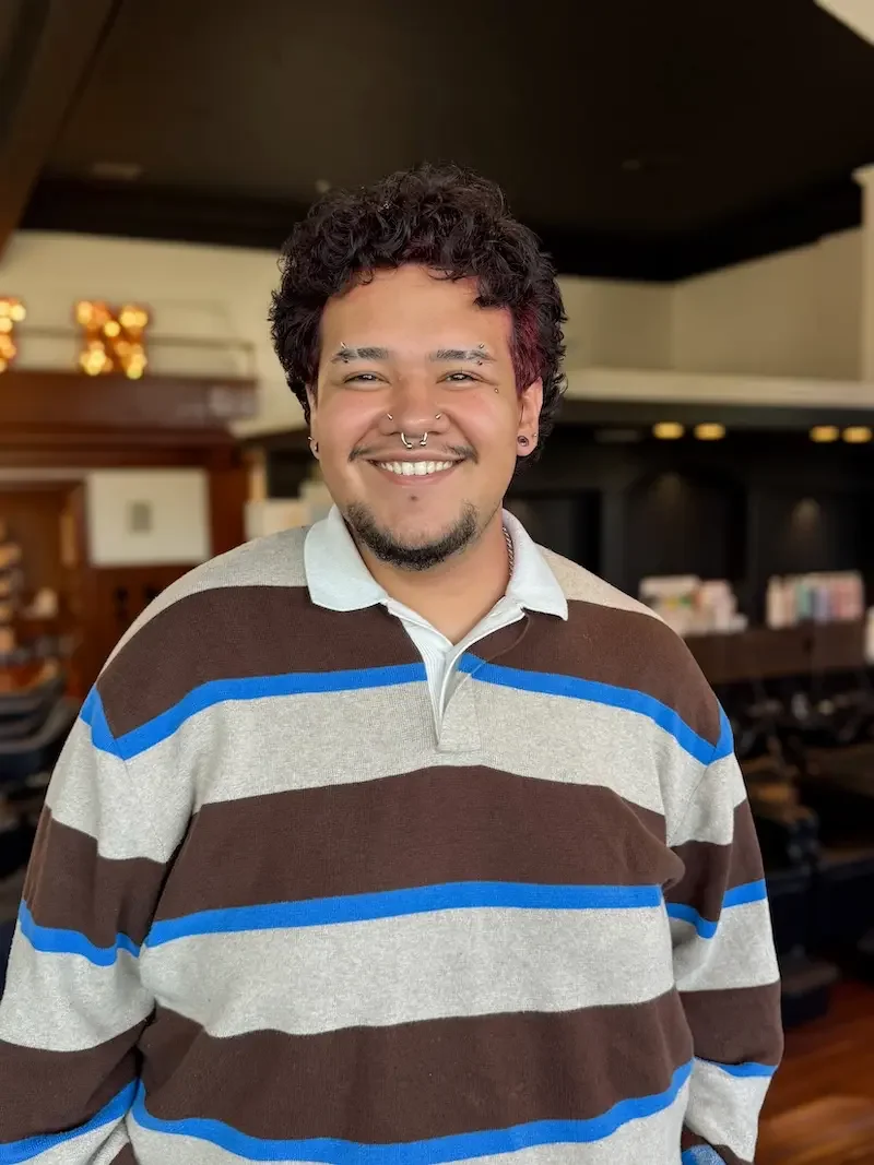 A person with short black curly hair wearing bold blue eyeshadow, dark lipstick, silver earrings, and a silver chain necklace, smiling indoors with decorative lighting in the background.