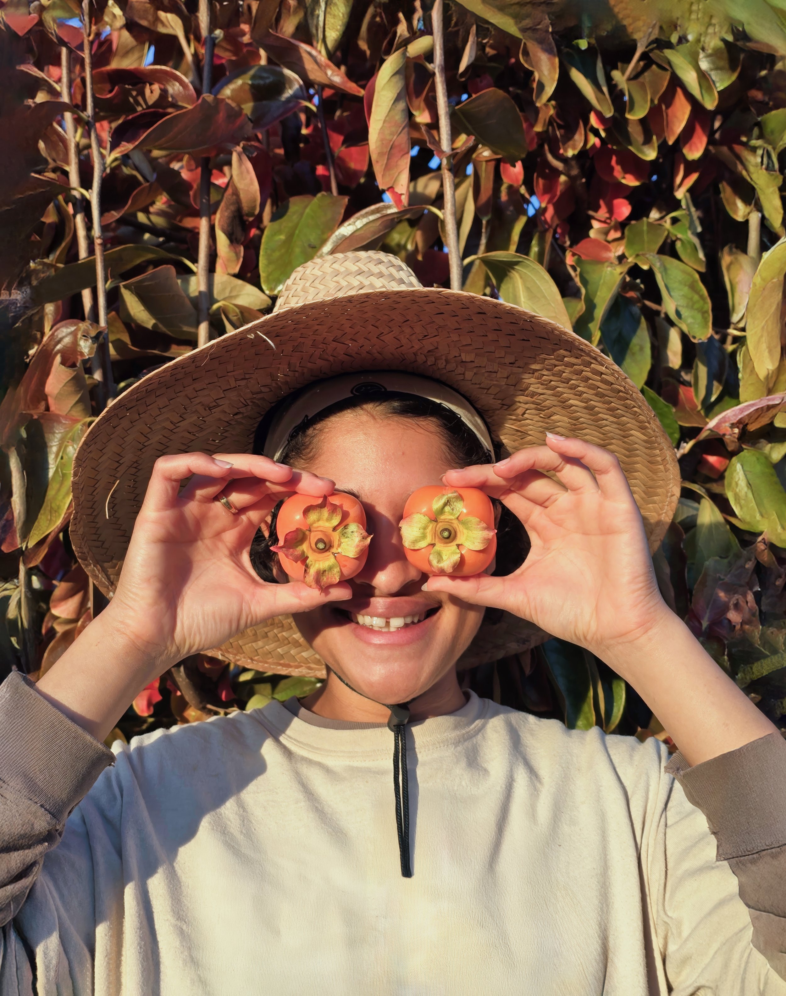 Kathryn stands in front of two persimmon trees holding two of the harvested fruits in front of her eyes like binoculars.