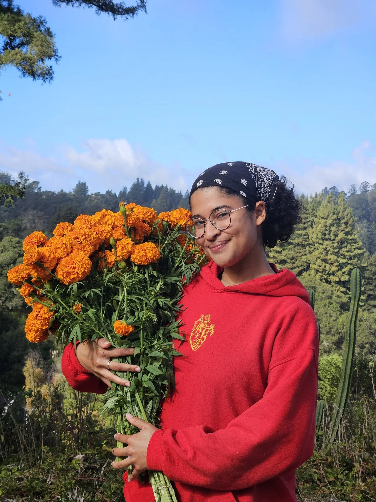 Kathryn holds a bundle or orange cempasuchiles and wear a read hoodie with a yellow heart embroidered on it, glasses and a black bandana.